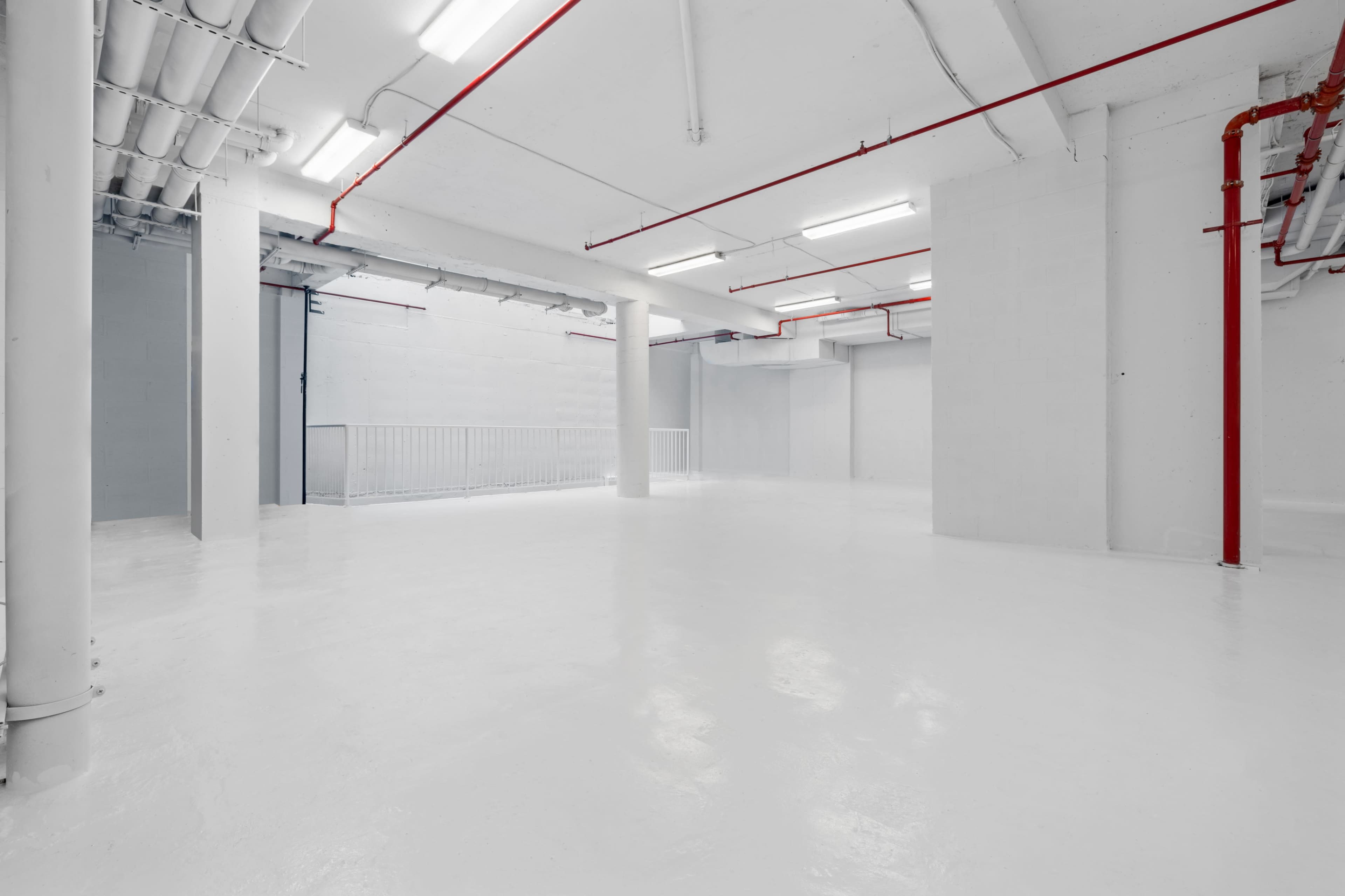 A large, empty industrial space with white walls and floors, illuminated by fluorescent lighting, featuring exposed pipes and minimal structural elements.