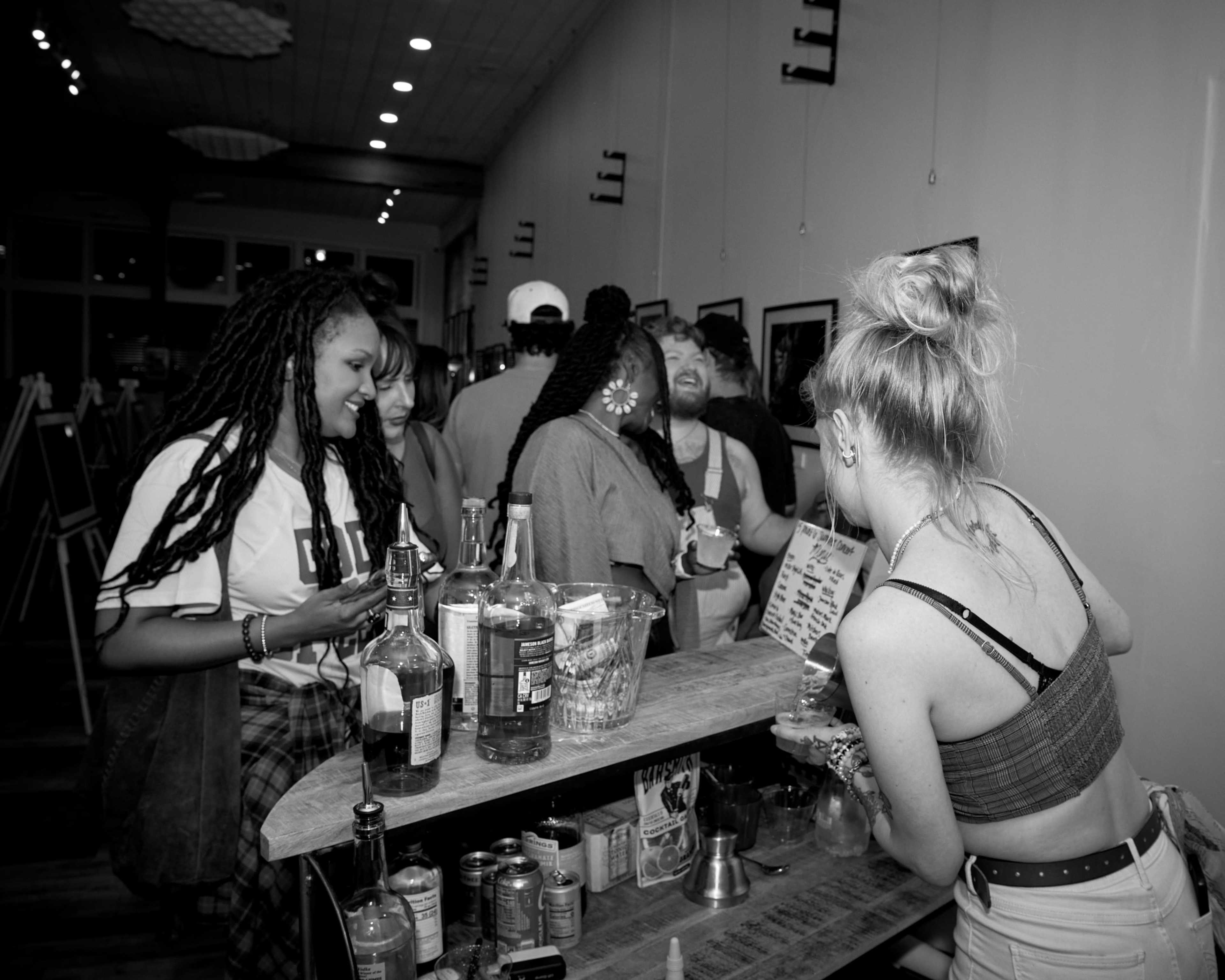 A group of people is gathered around a bar, with some engaged in conversation and others ordering drinks.