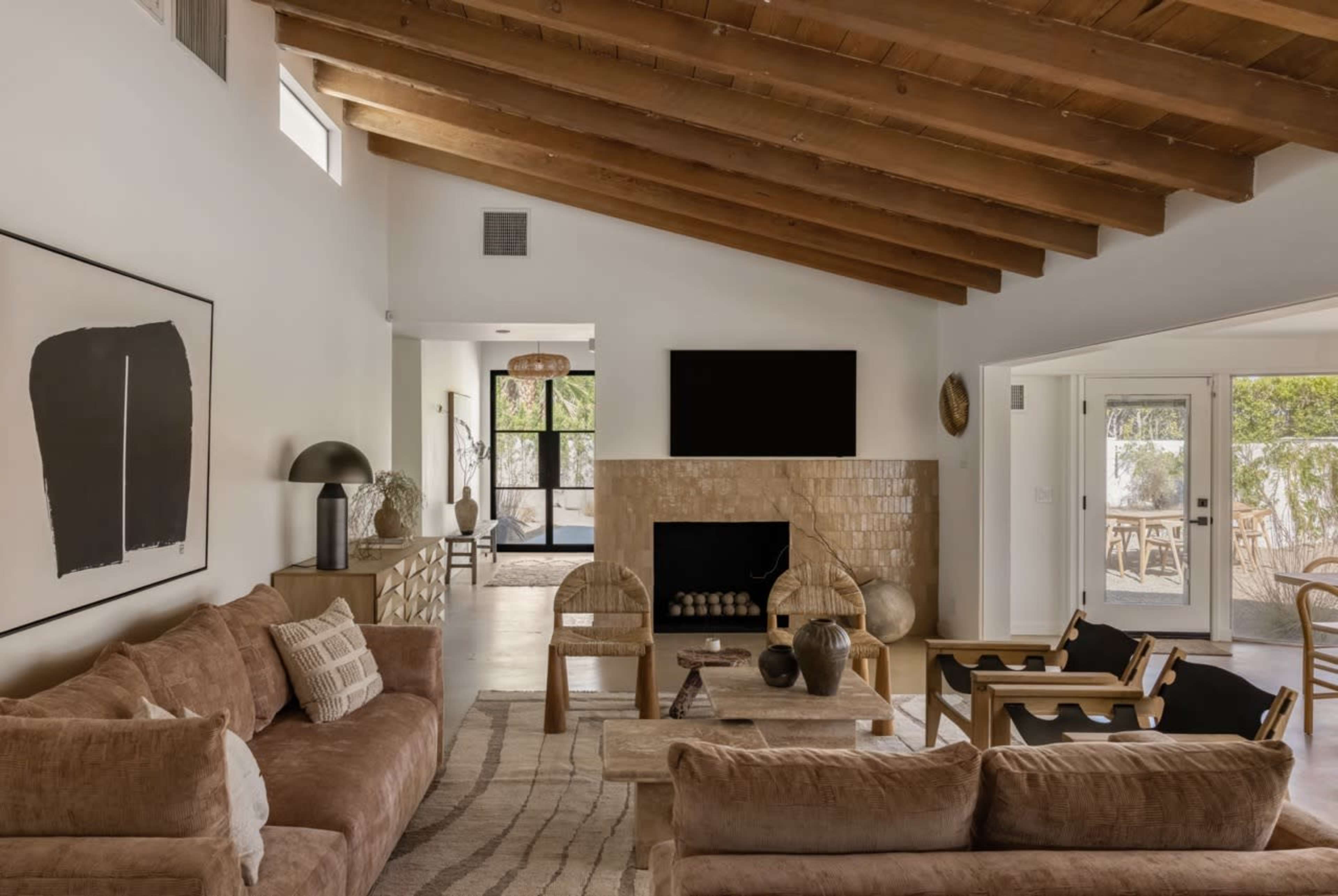 A spacious living room features a beige sectional sofa, wooden beams on the ceiling, and a fireplace with a tiled facade, leading to an outdoor area through large sliding glass doors.