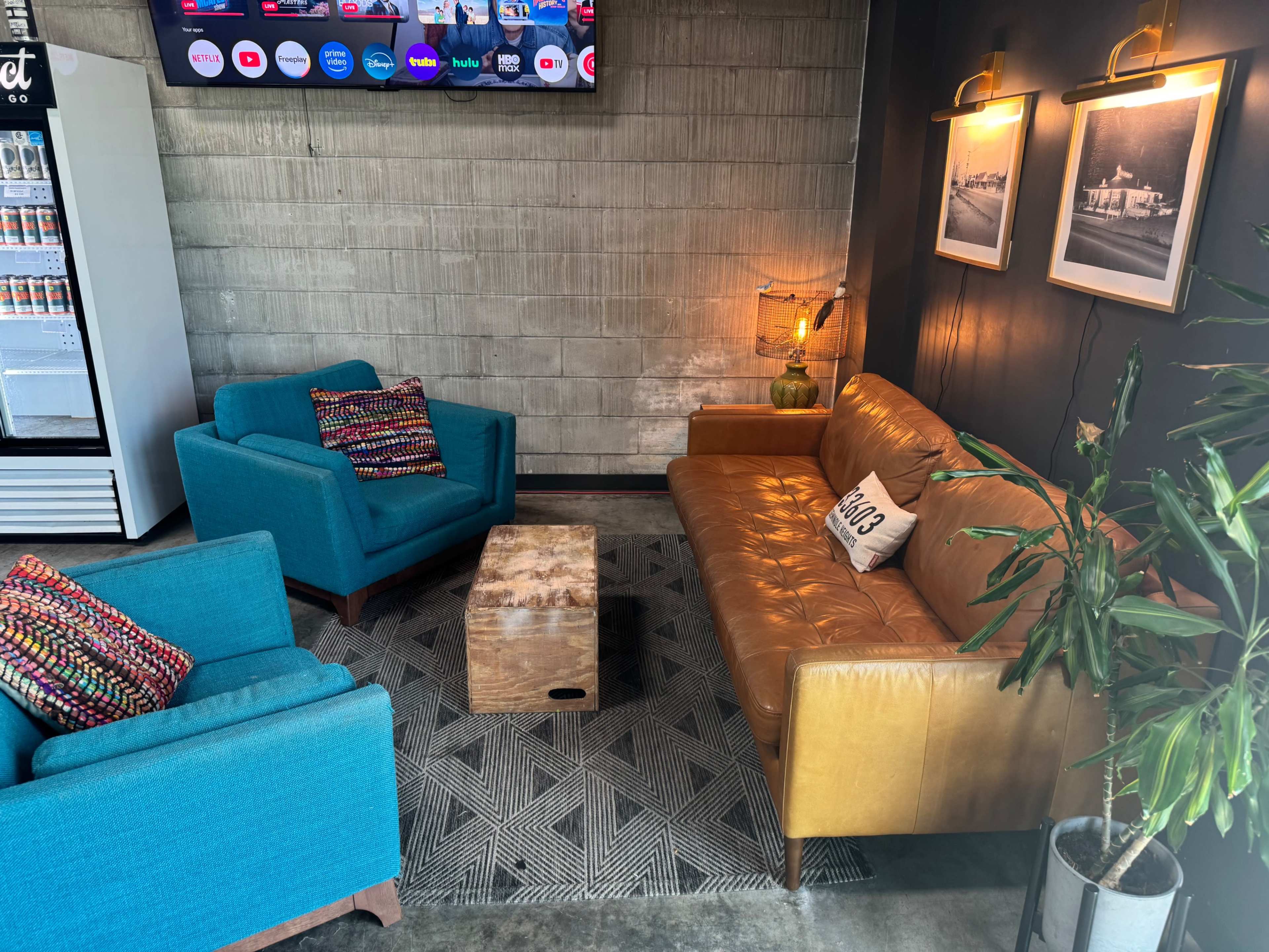 The image shows a cozy seating area with two colorful armchairs, a brown leather sofa, a wooden coffee table, and wall-mounted lights, all set against a textured gray wall.