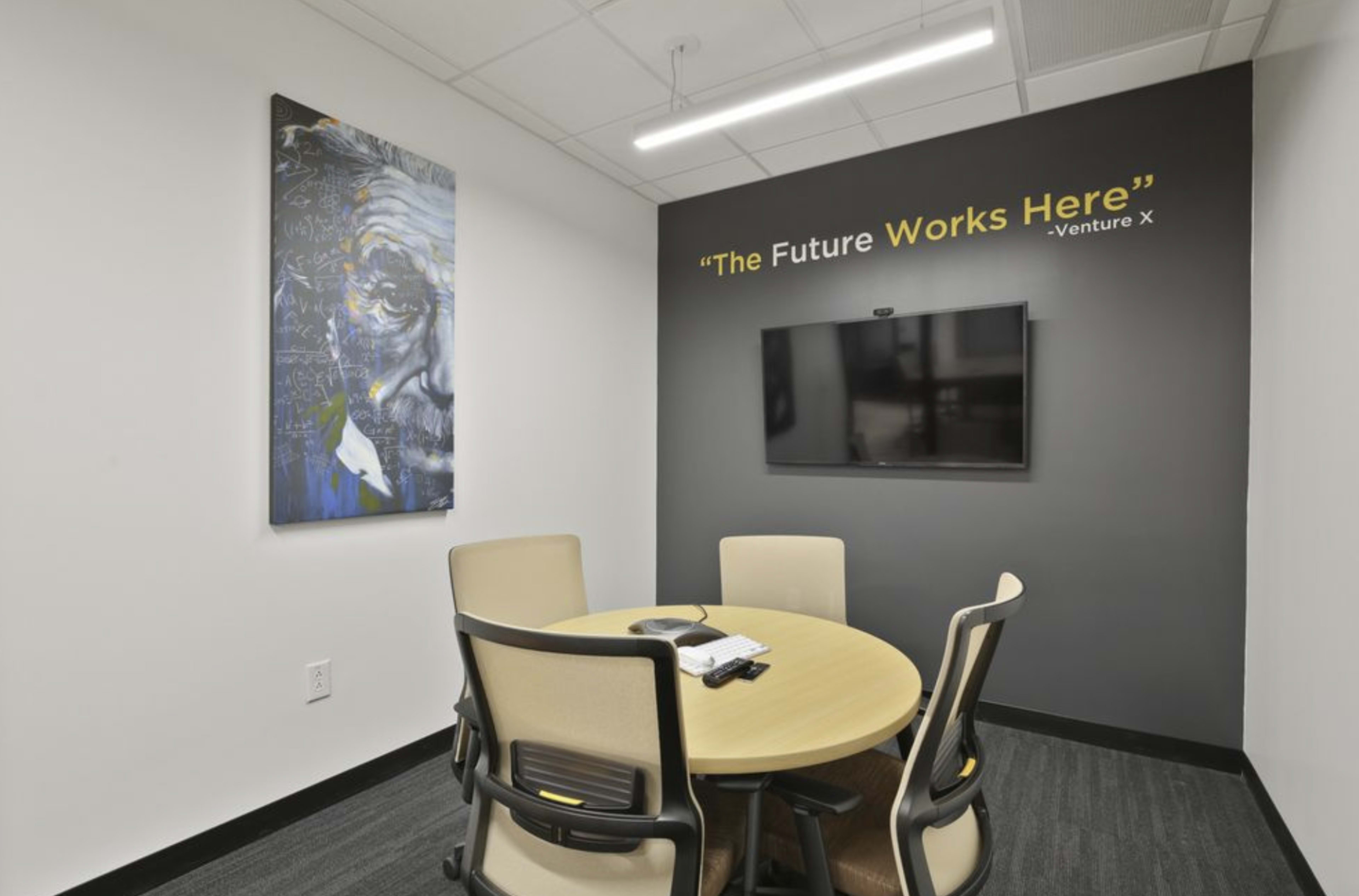 A small conference room features a round table with four chairs, a wall-mounted television, and a portrait of Albert Einstein, accompanied by the phrase "The Future Works Here" painted on the wall.