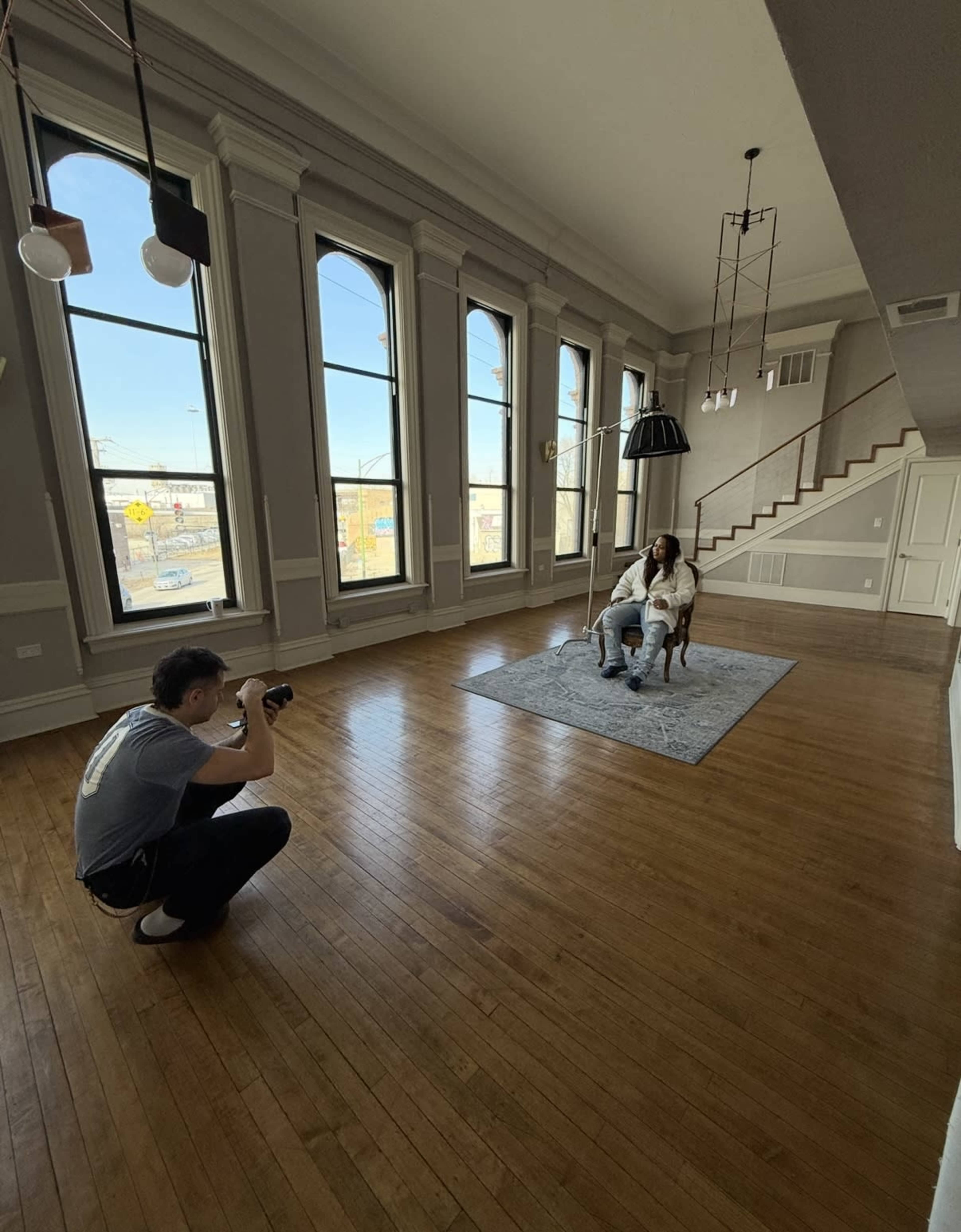 A person is taking a photograph of another person sitting on a chair in a spacious, well-lit room with large windows and wooden floors.