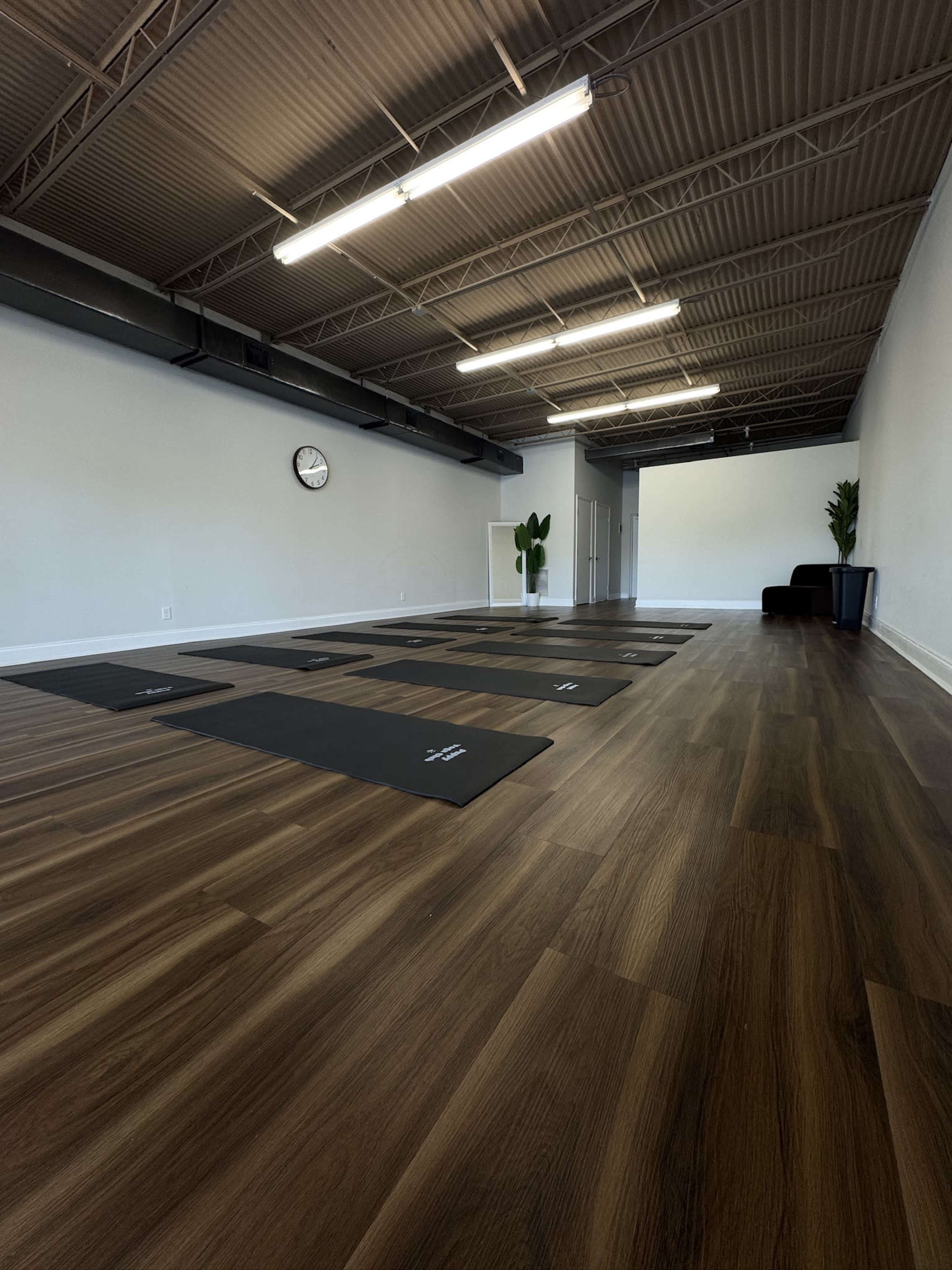 The image shows a spacious yoga studio with several yoga mats arranged on a polished wooden floor, under bright overhead lighting.