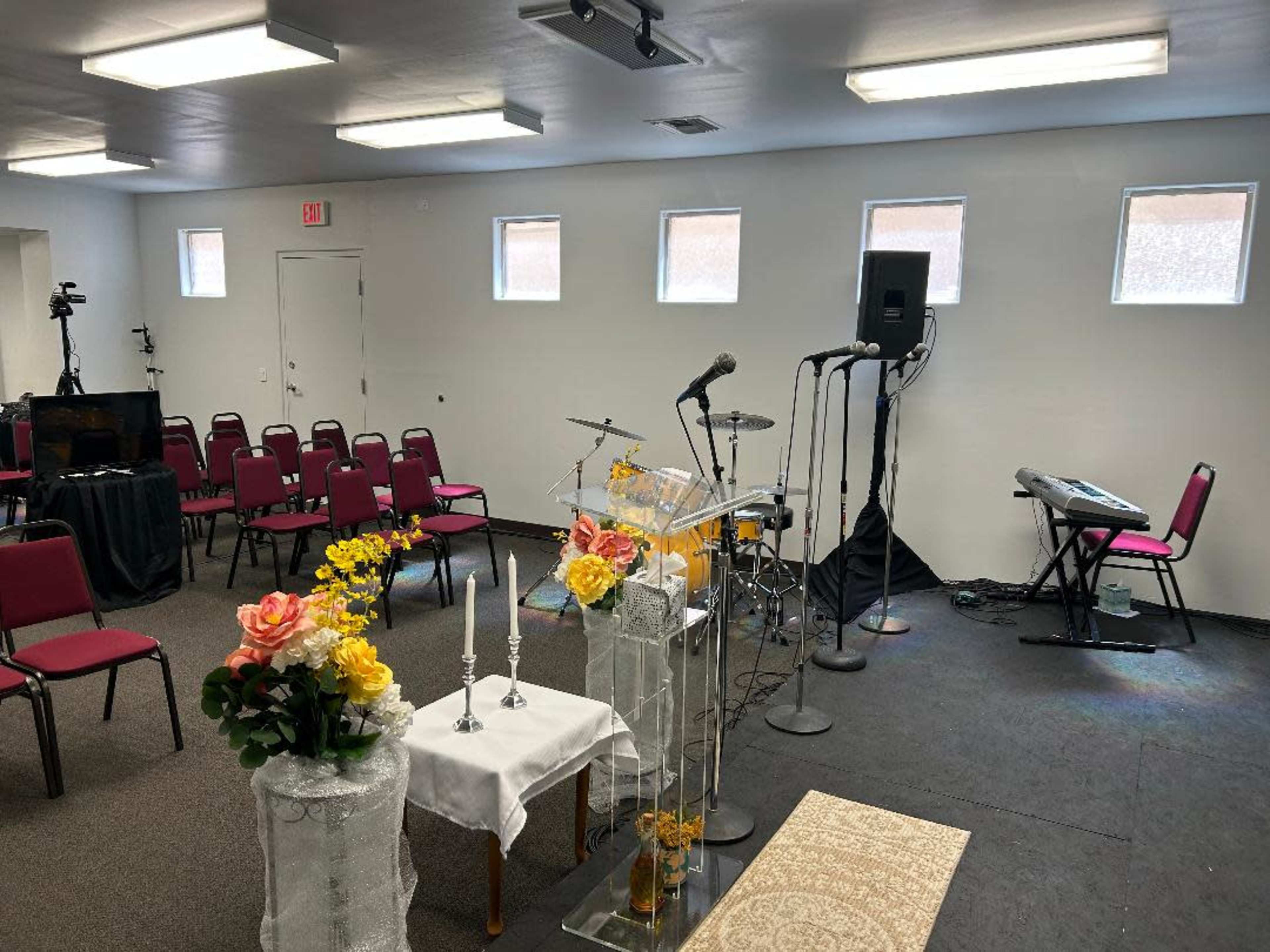 A small room arranged with chairs for an event, featuring a stage setup with microphones, musical instruments, and decorative floral arrangements.