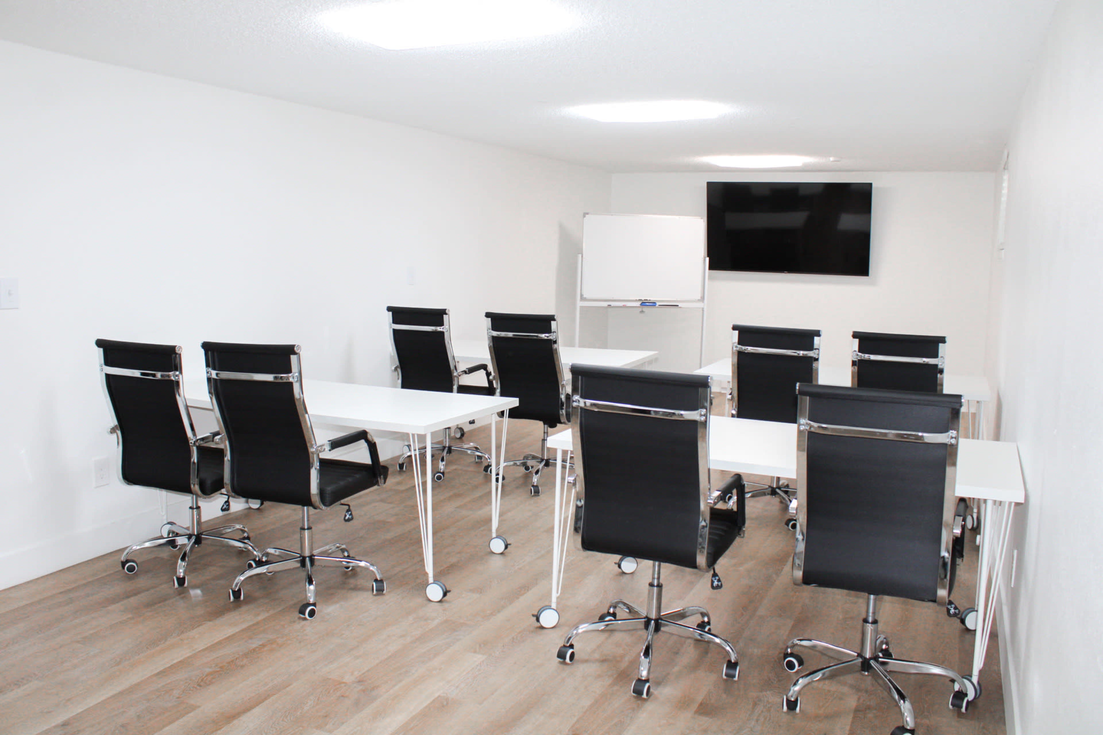 A minimalist conference room features several black rolling chairs around white tables, with a whiteboard and a television mounted on the wall.