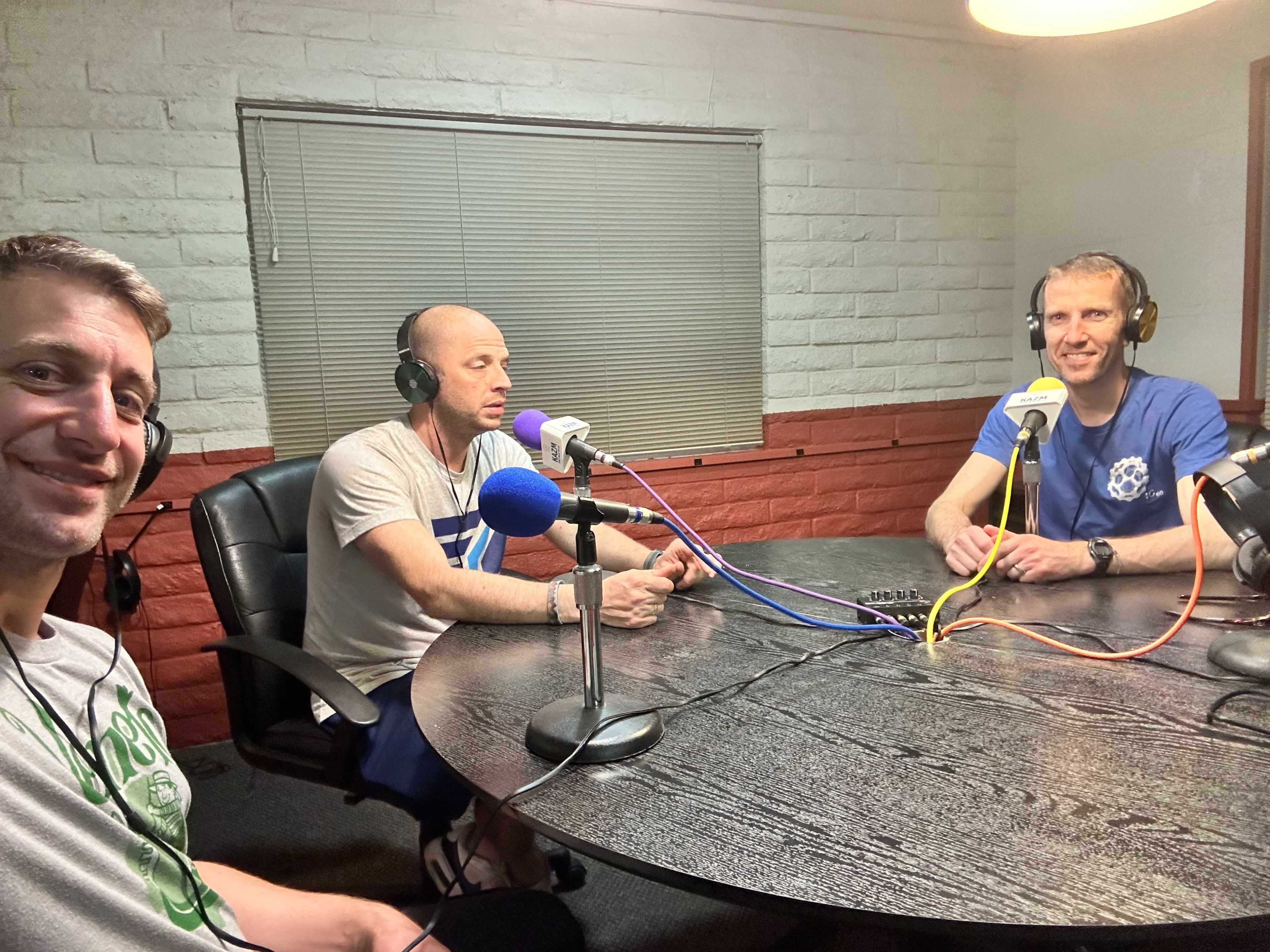 Three men are sitting at a table in a recording studio, wearing headphones and microphones, as they engage in a conversation.