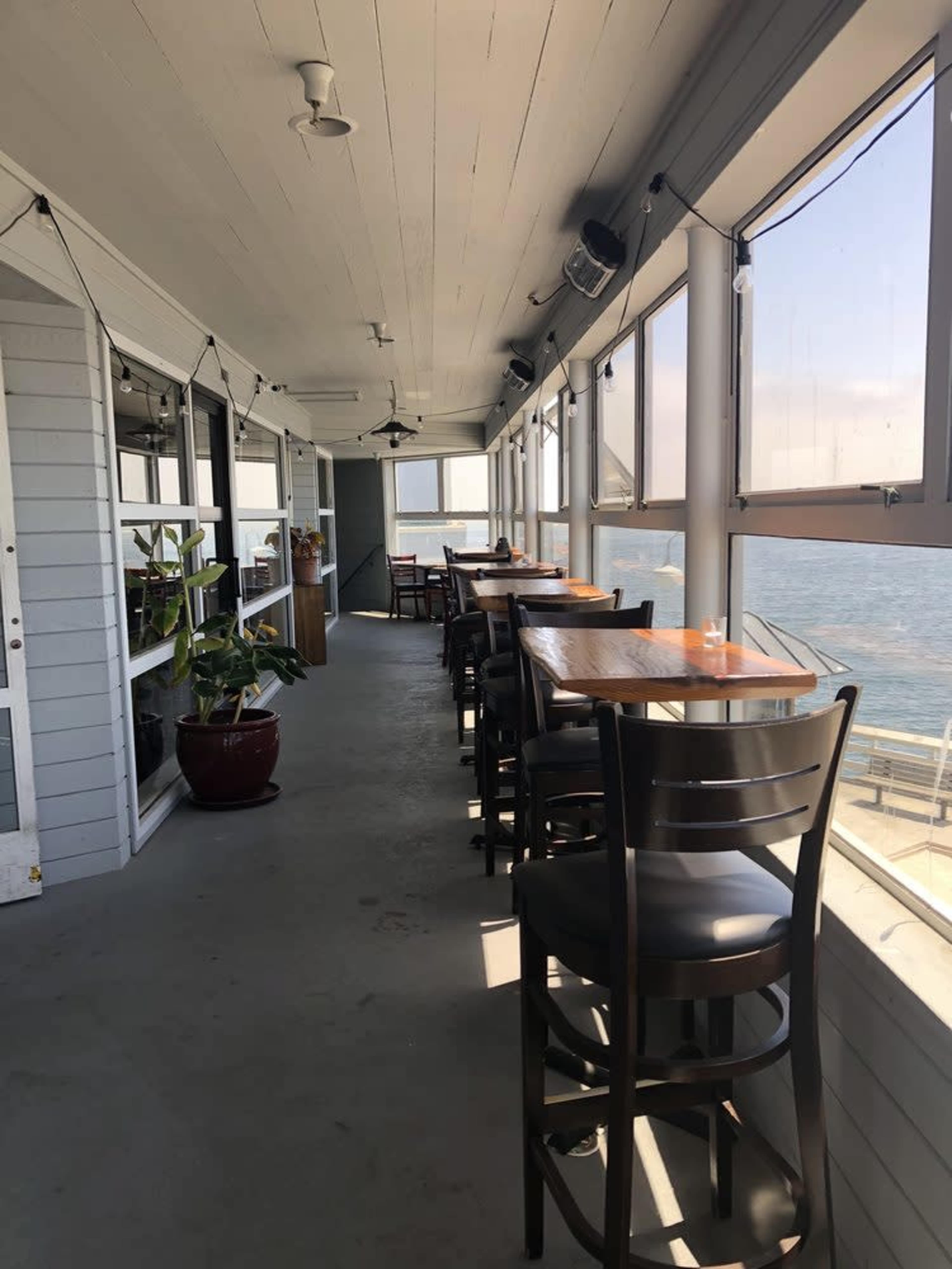 The image shows a narrow, sunlit passageway with several tall tables and chairs lining the sides, overlooking a body of water through large windows.