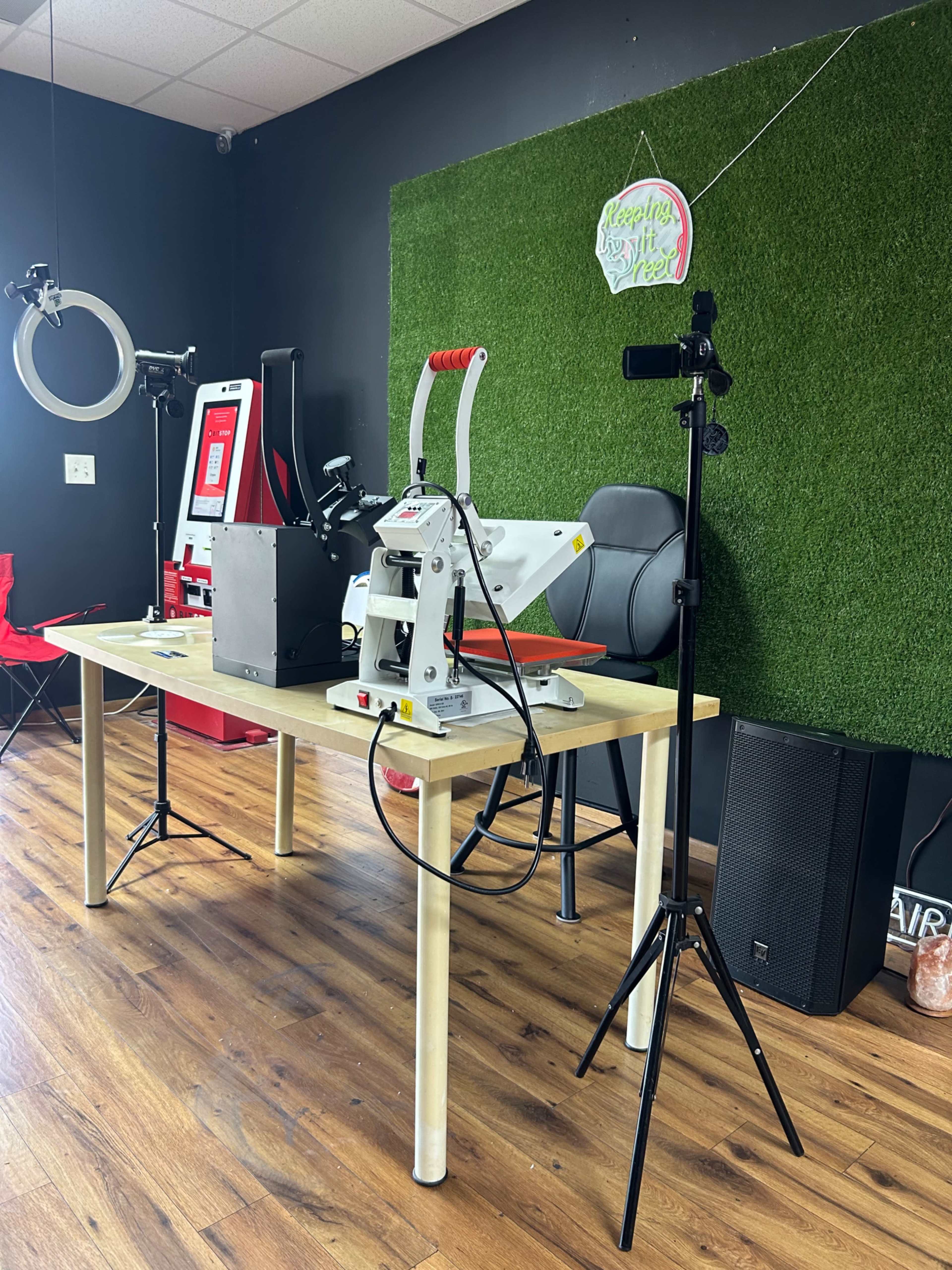 The image shows a workspace with a heat press machine on a table, surrounded by a green felt wall, various equipment, and a microphone setup.