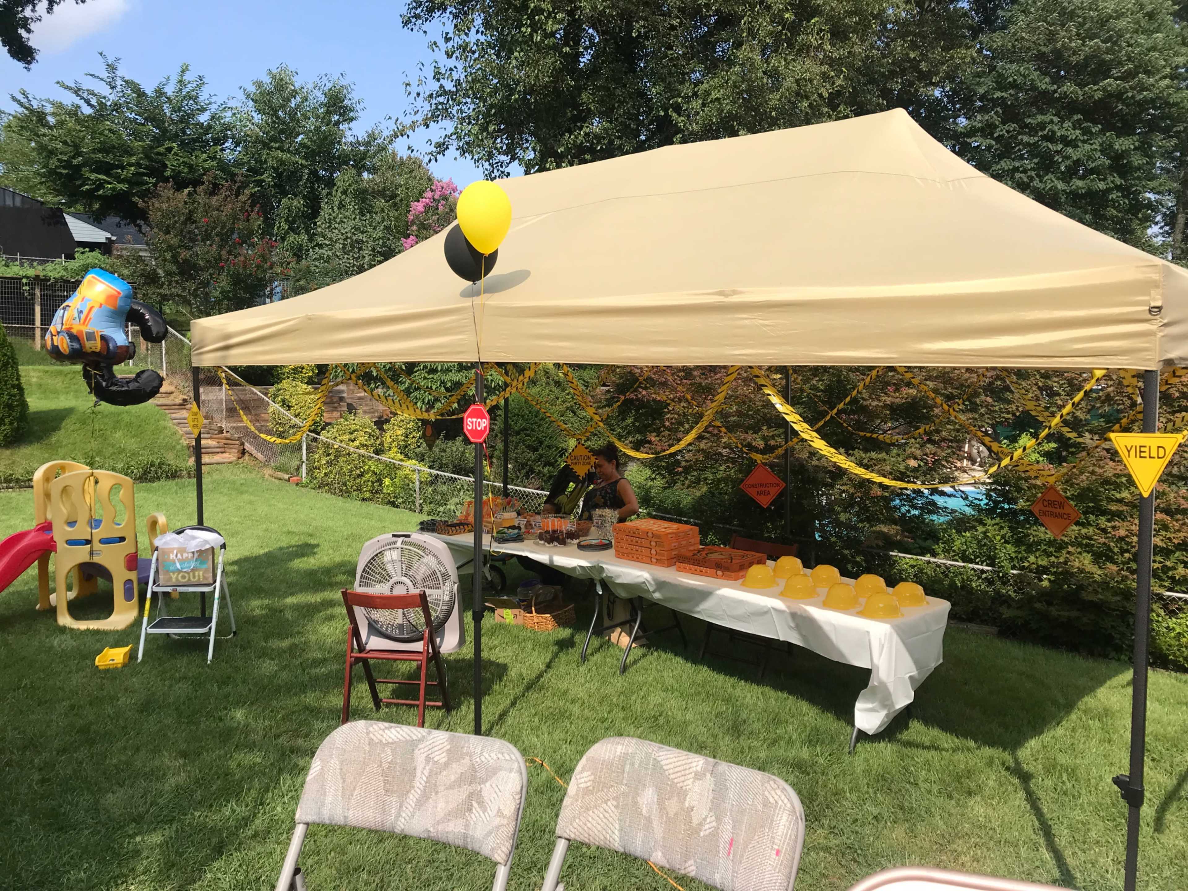 A large tent is set up in a grassy area, with a table displaying yellow balloons and treats, surrounded by chairs and decorations.