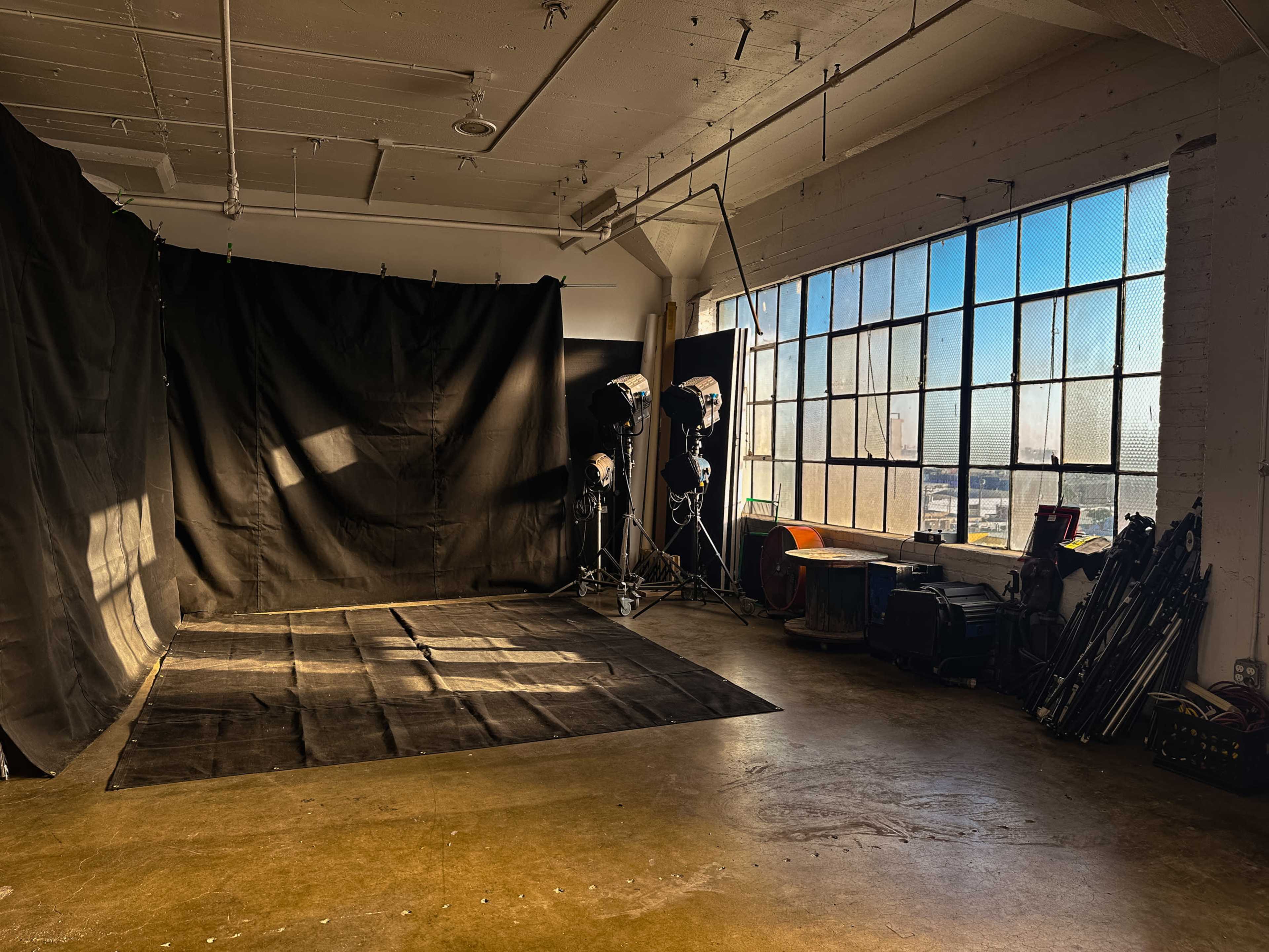 The image shows a spacious photography studio with a large window, black backdrop, lighting equipment, and various studio accessories along the walls.