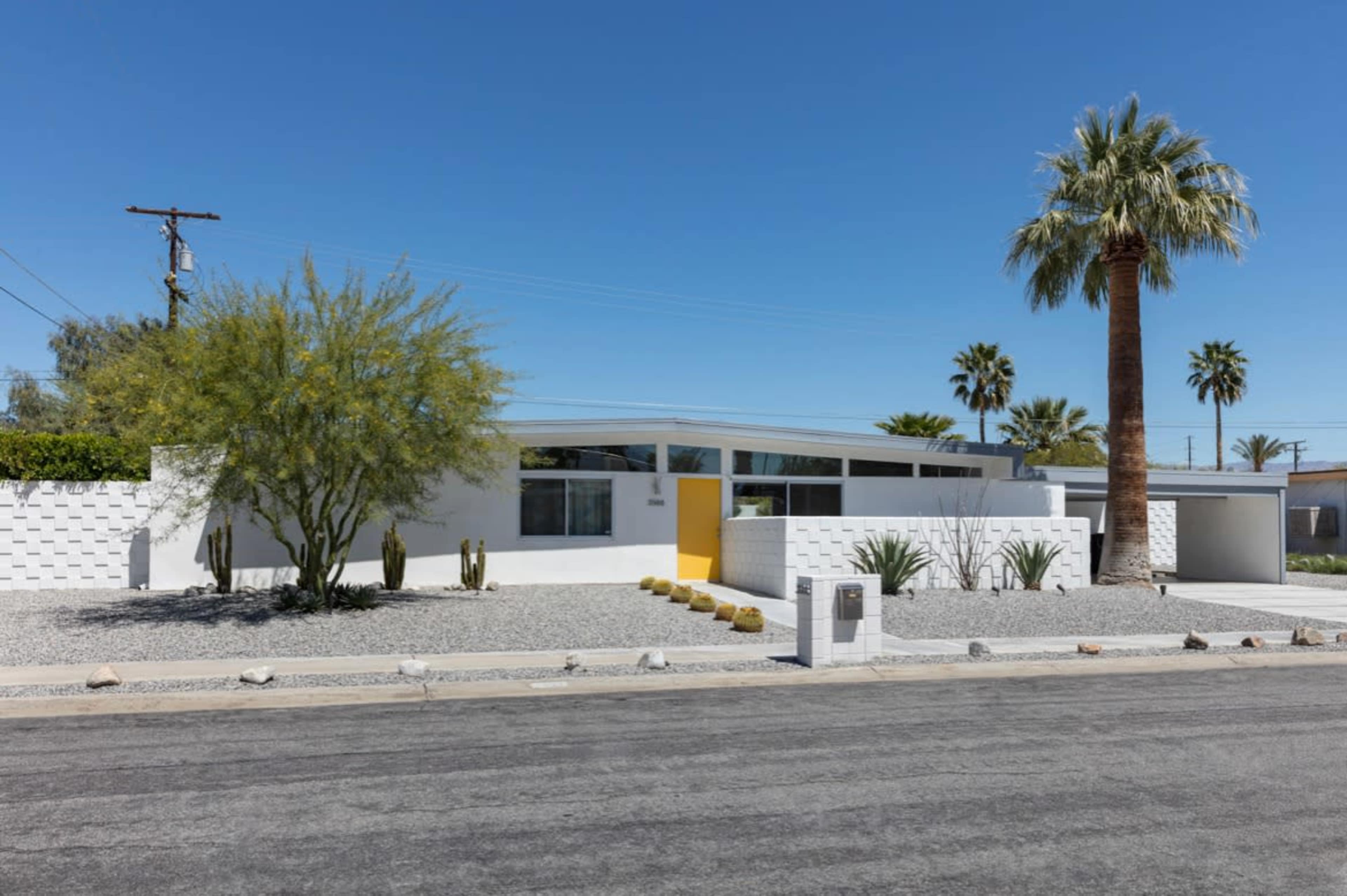 The image shows a modern, single-story house with a white exterior, yellow front door, and landscaped desert vegetation.