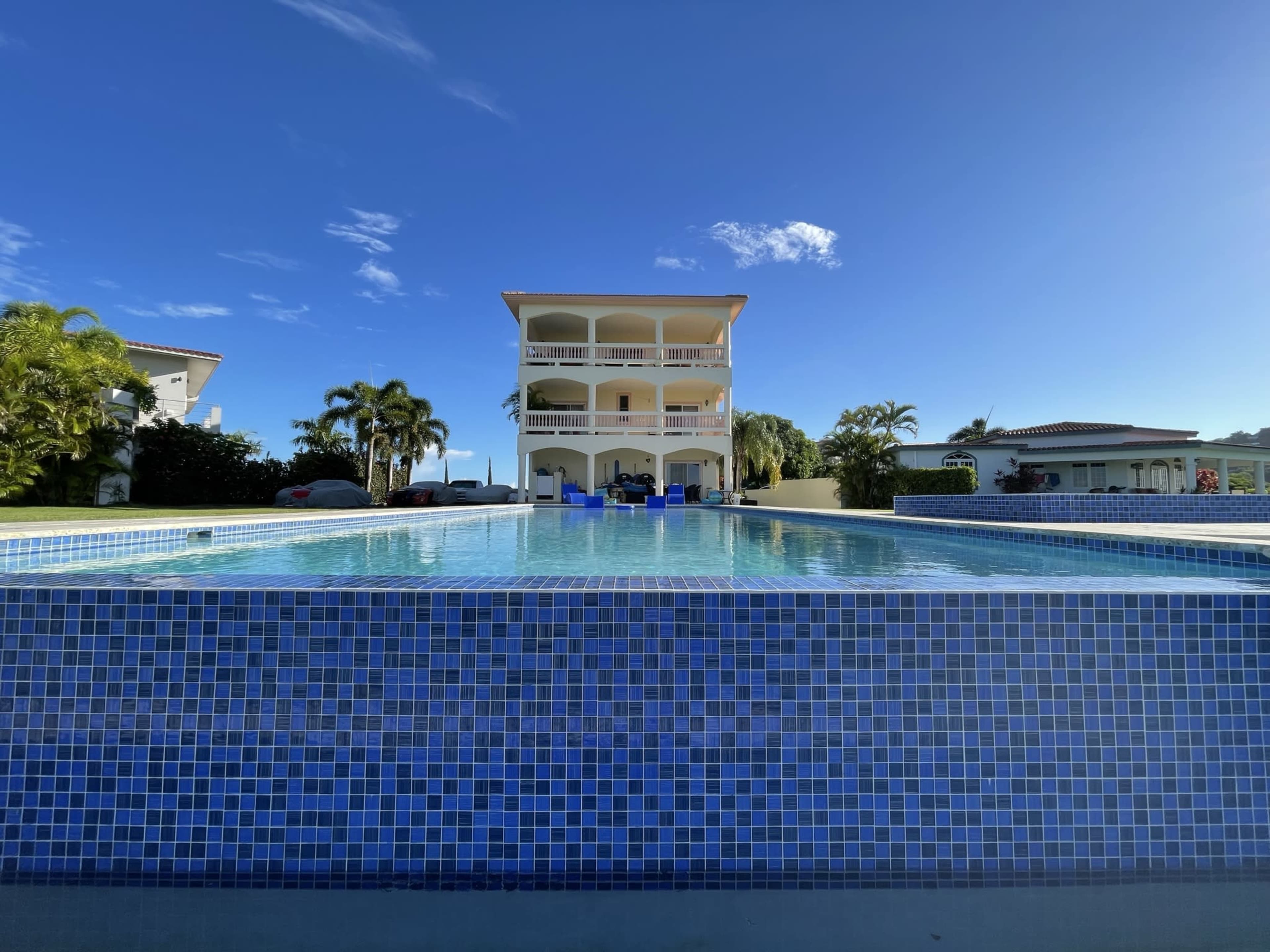 A three-story building overlooks a swimming pool with a mosaic blue tile design under a clear blue sky.