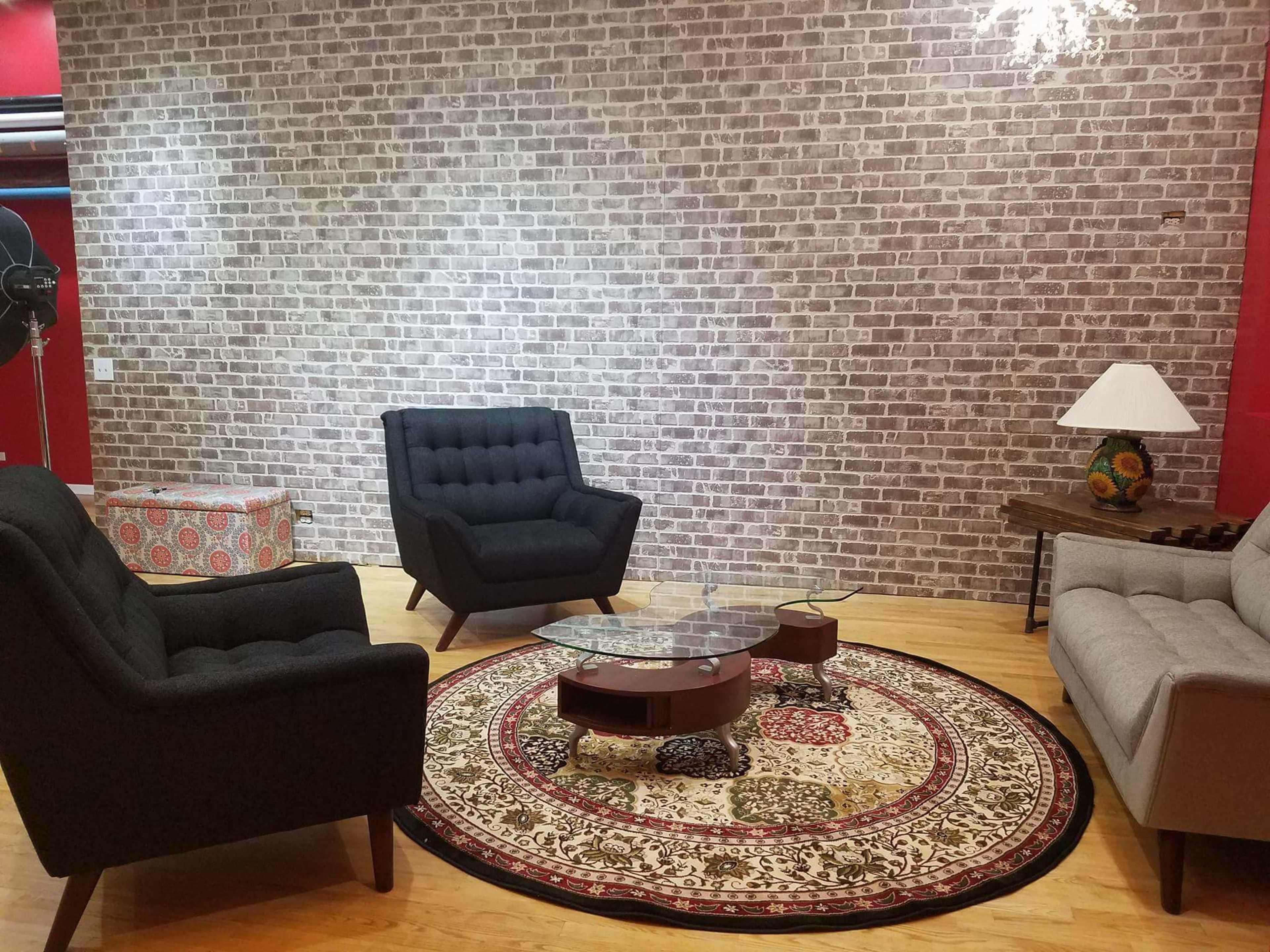A modern living room with two black armchairs, a light gray sofa, a round coffee table, and a patterned area rug set against a brick wall.