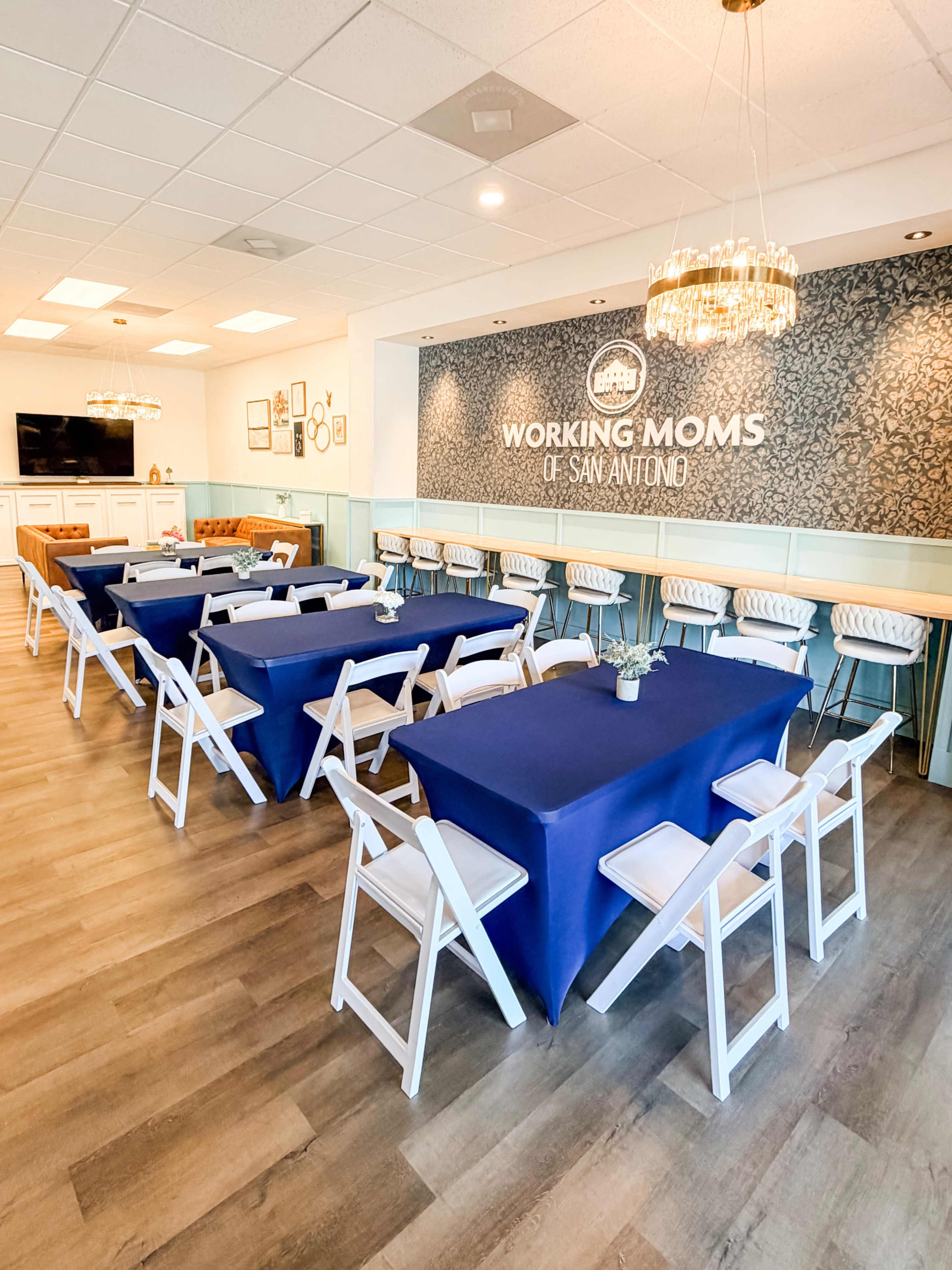 The image shows a well-organized meeting room with navy blue tablecloths covering rectangular tables, white chairs, and a decorative wall that reads "Working Moms of San Antonio."
