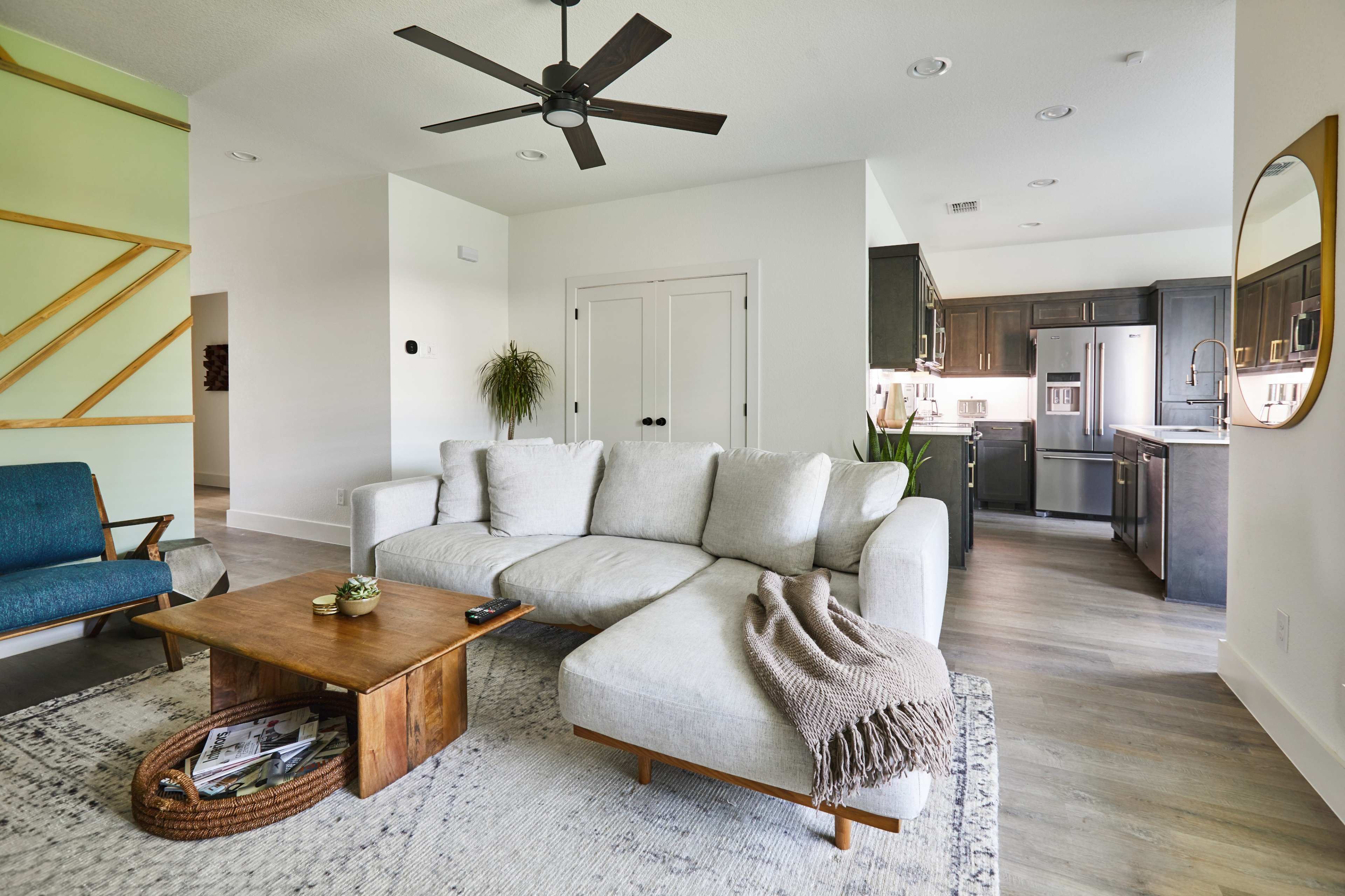 The image shows a modern living room with a light gray sectional sofa, a wooden coffee table, and a kitchen visible in the background.