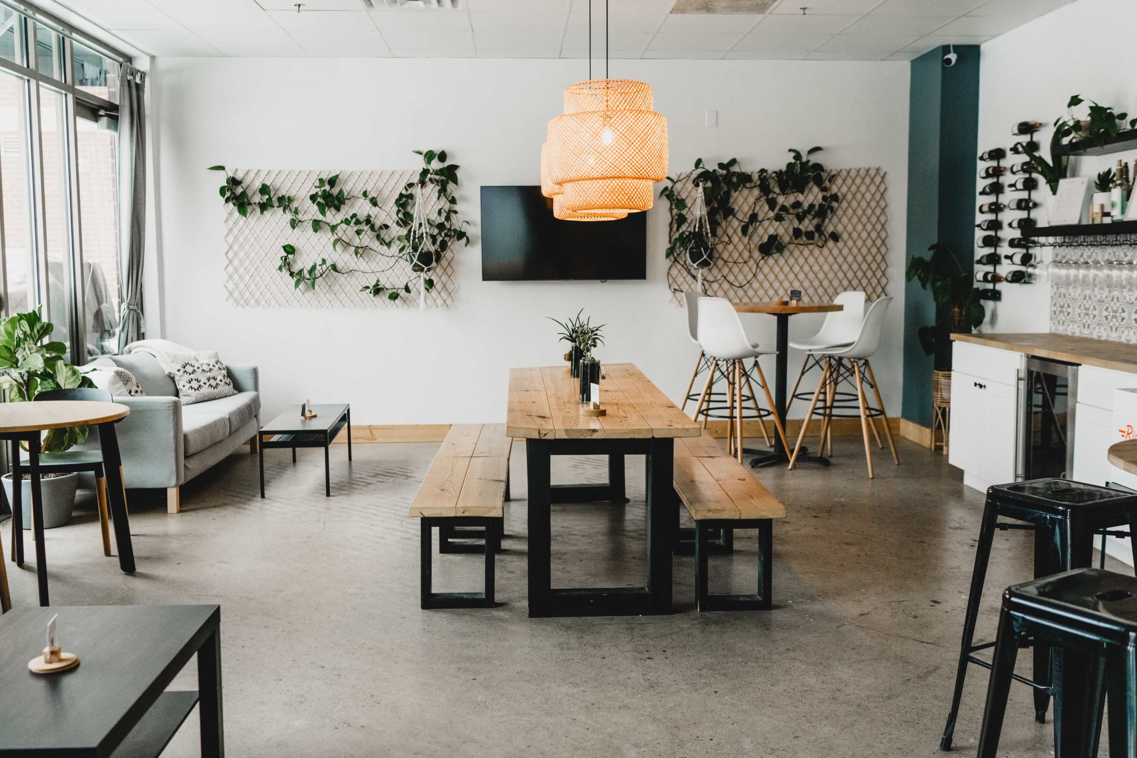 The image shows a modern café interior with wooden tables, a large pendant light, and greenery on the walls.