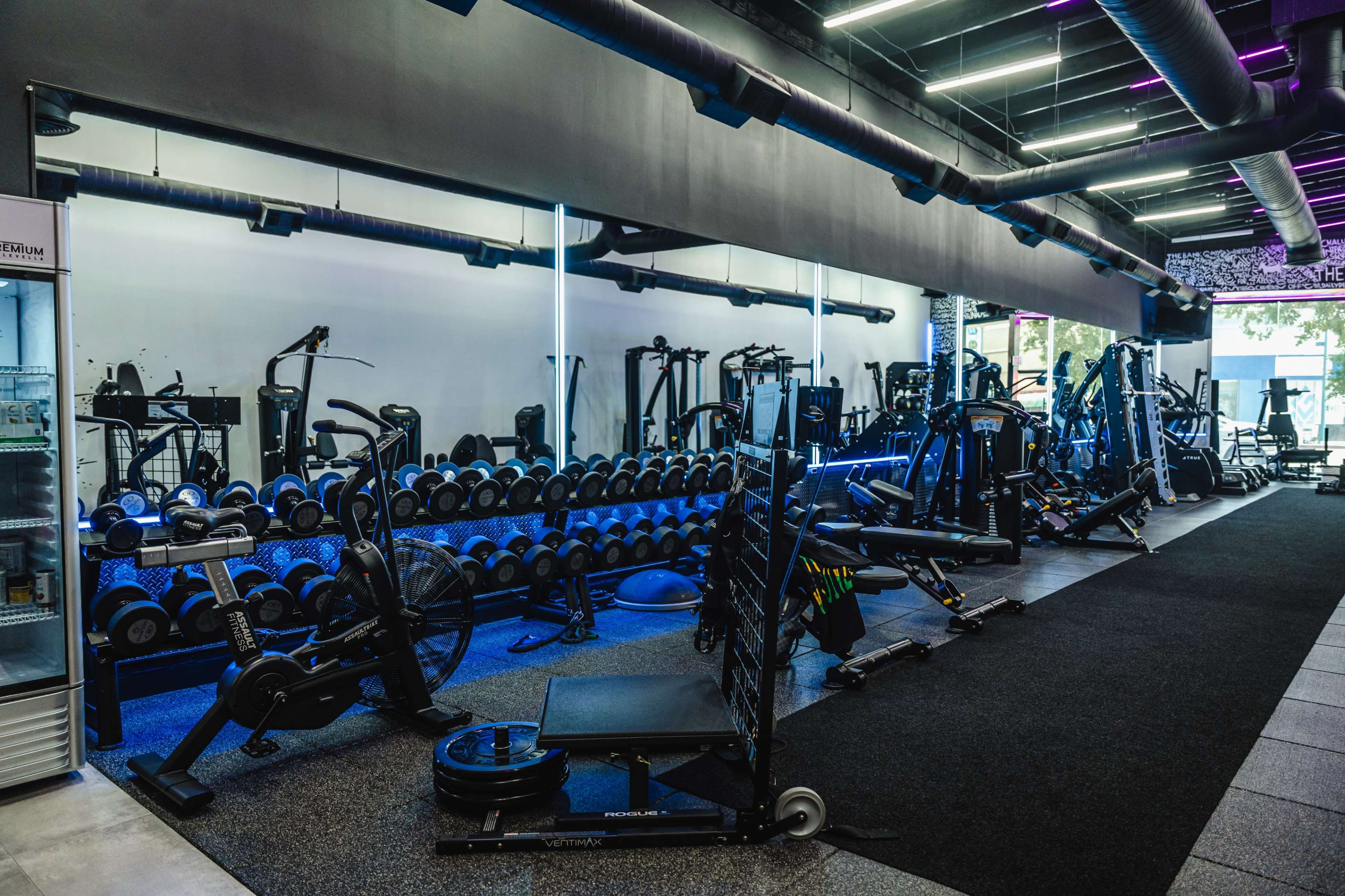 The image shows an indoor gym equipped with various exercise machines, free weights, and fitness equipment, organized along a spacious area with dark flooring and neon lighting.