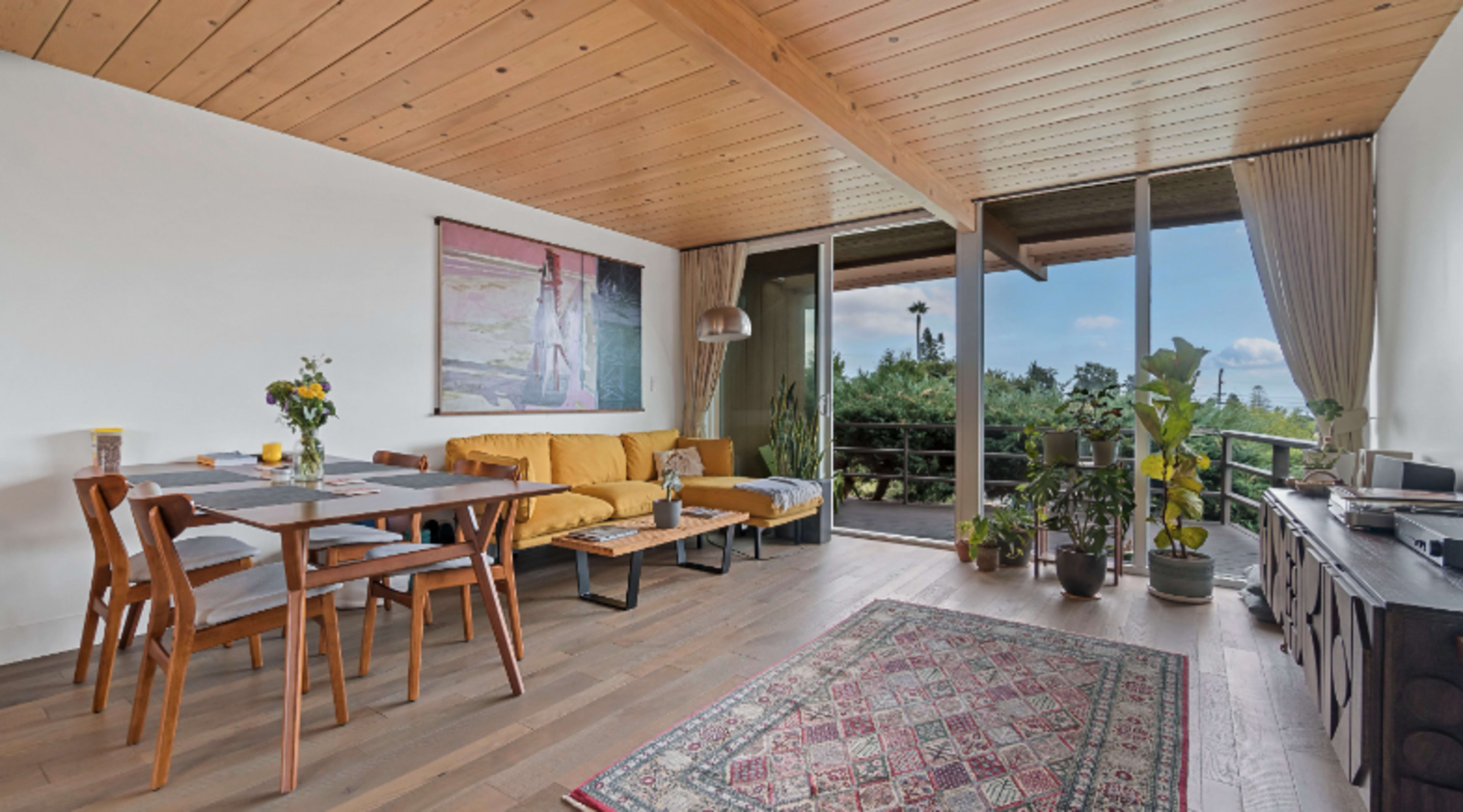 A modern living space featuring a dining table near a sofa, large windows with views of greenery, and various plants around the room.