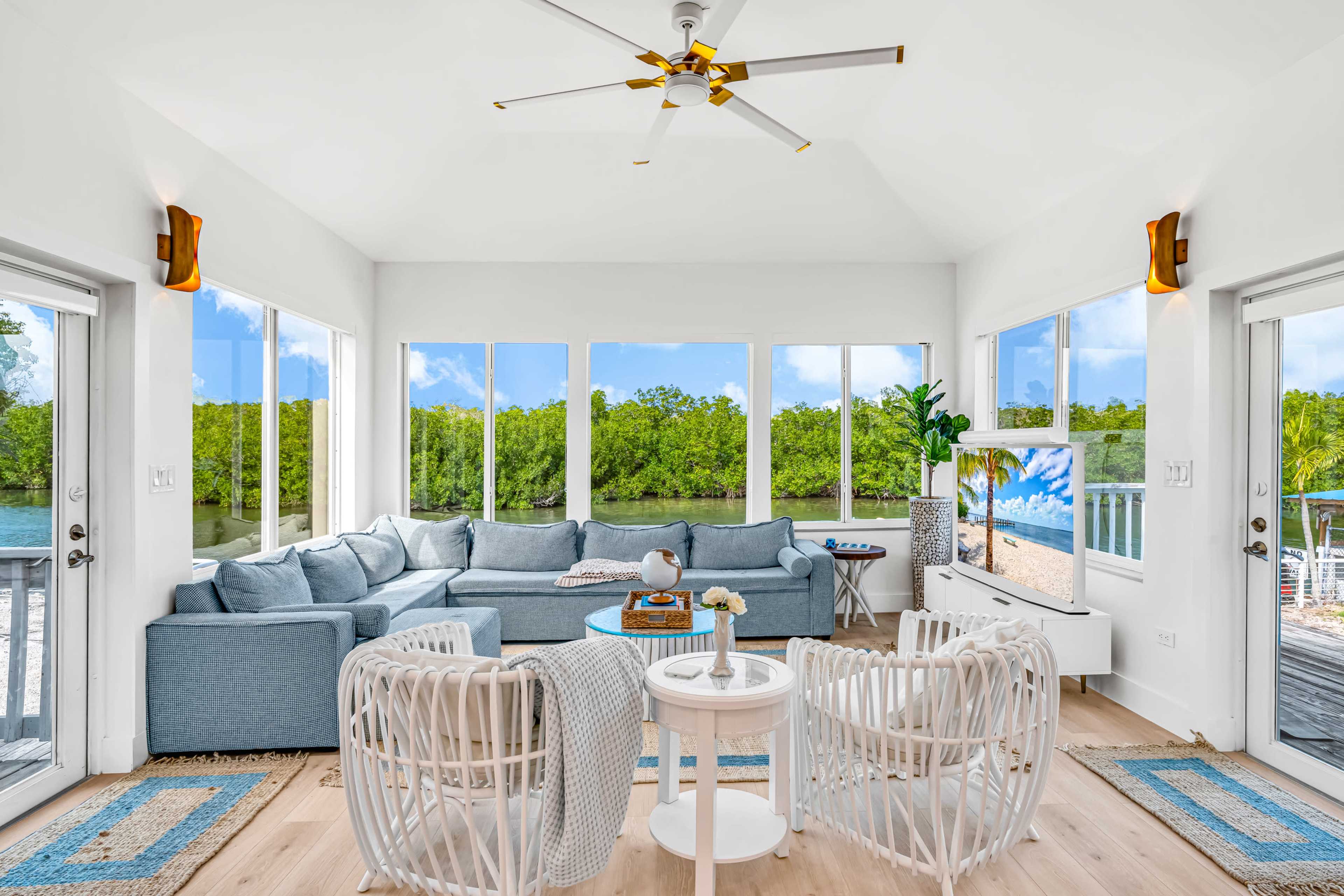 The image shows a bright, airy living room with large windows, a blue sectional sofa, and white accent furniture, overlooking a natural landscape.