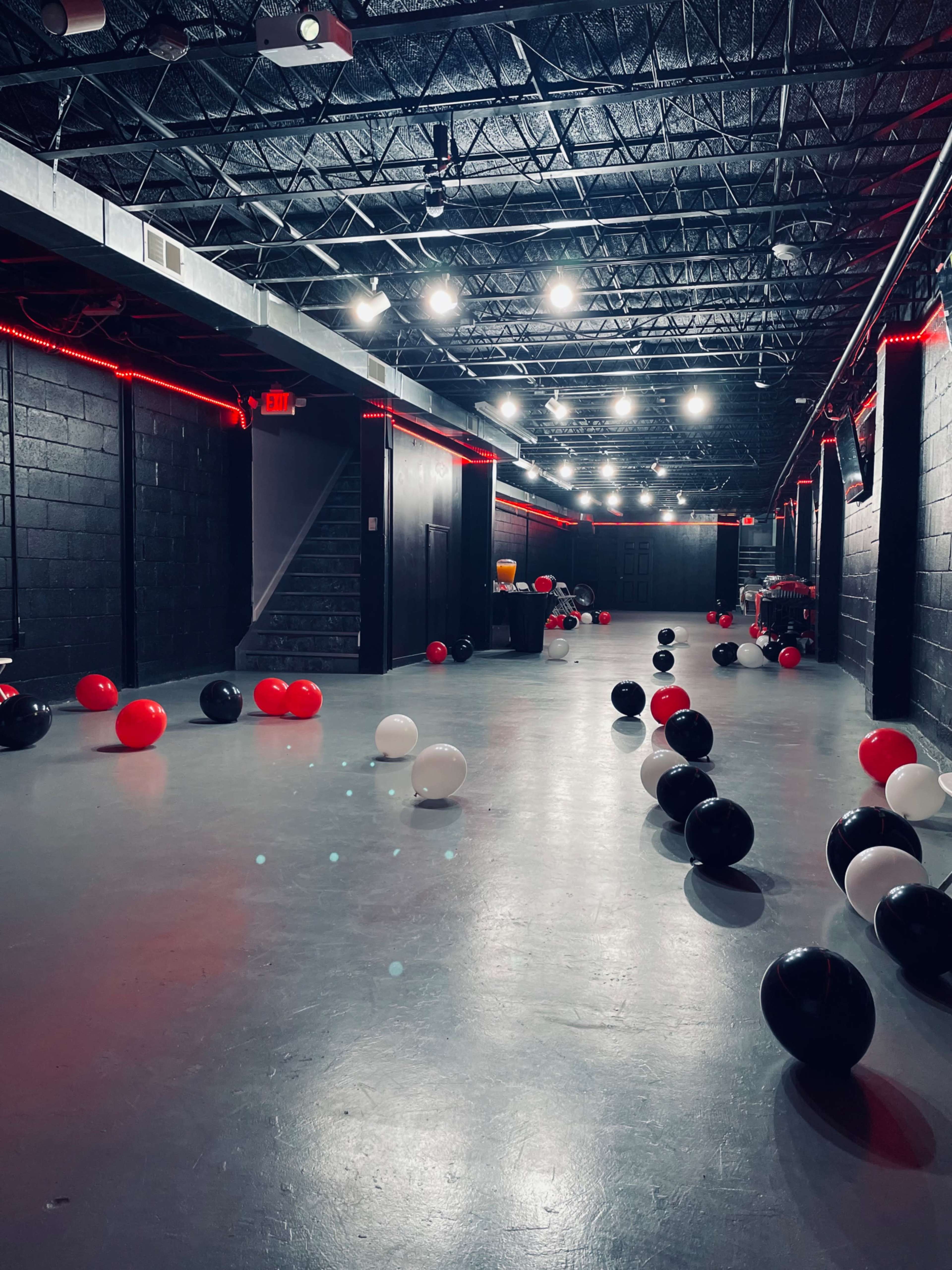 A dimly lit, empty space with black walls and a gray floor, scattered with red, black, and white balloons.