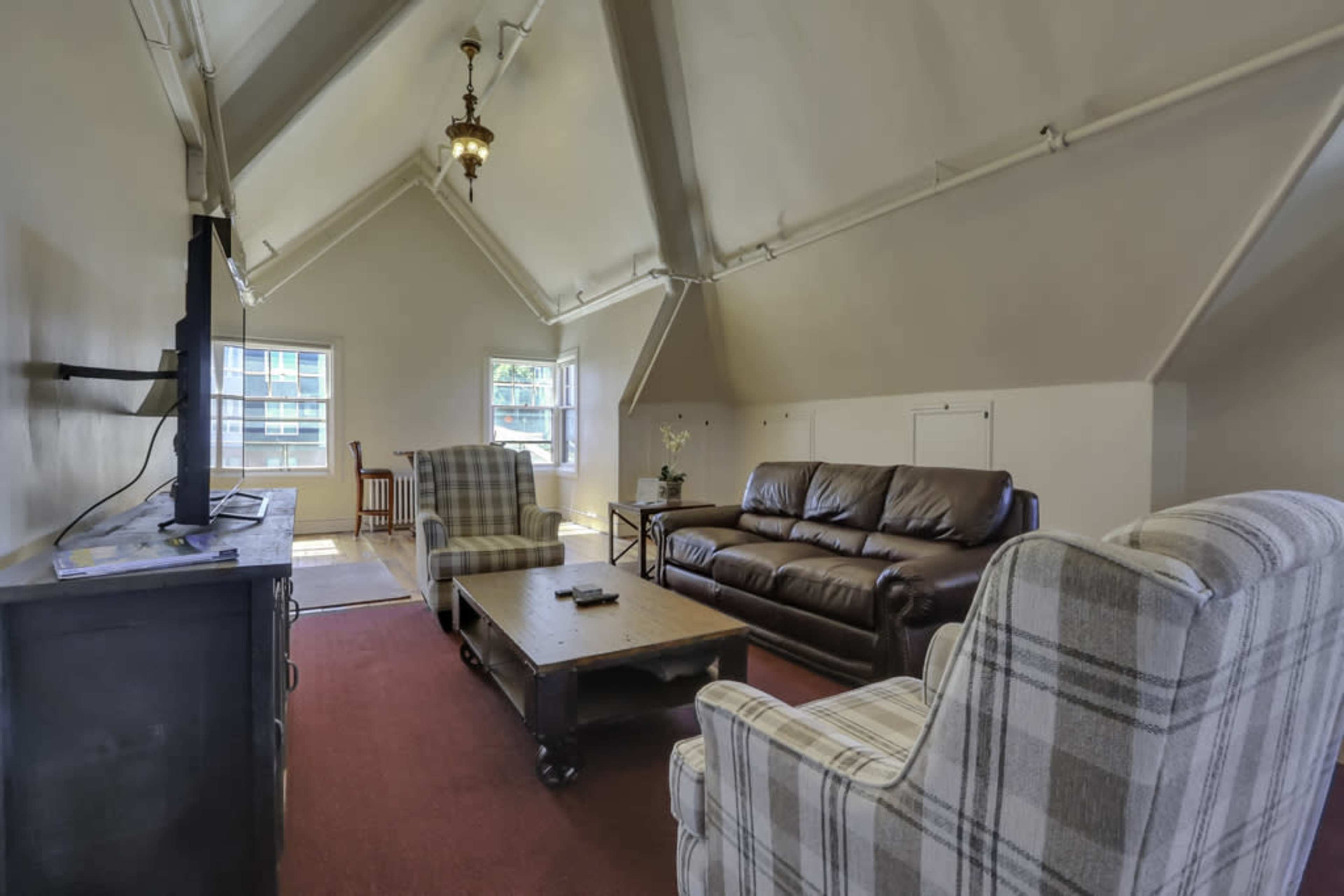 A spacious living room with a brown leather sofa, two plaid armchairs, and a wooden coffee table under a high ceiling with a decorative light fixture.