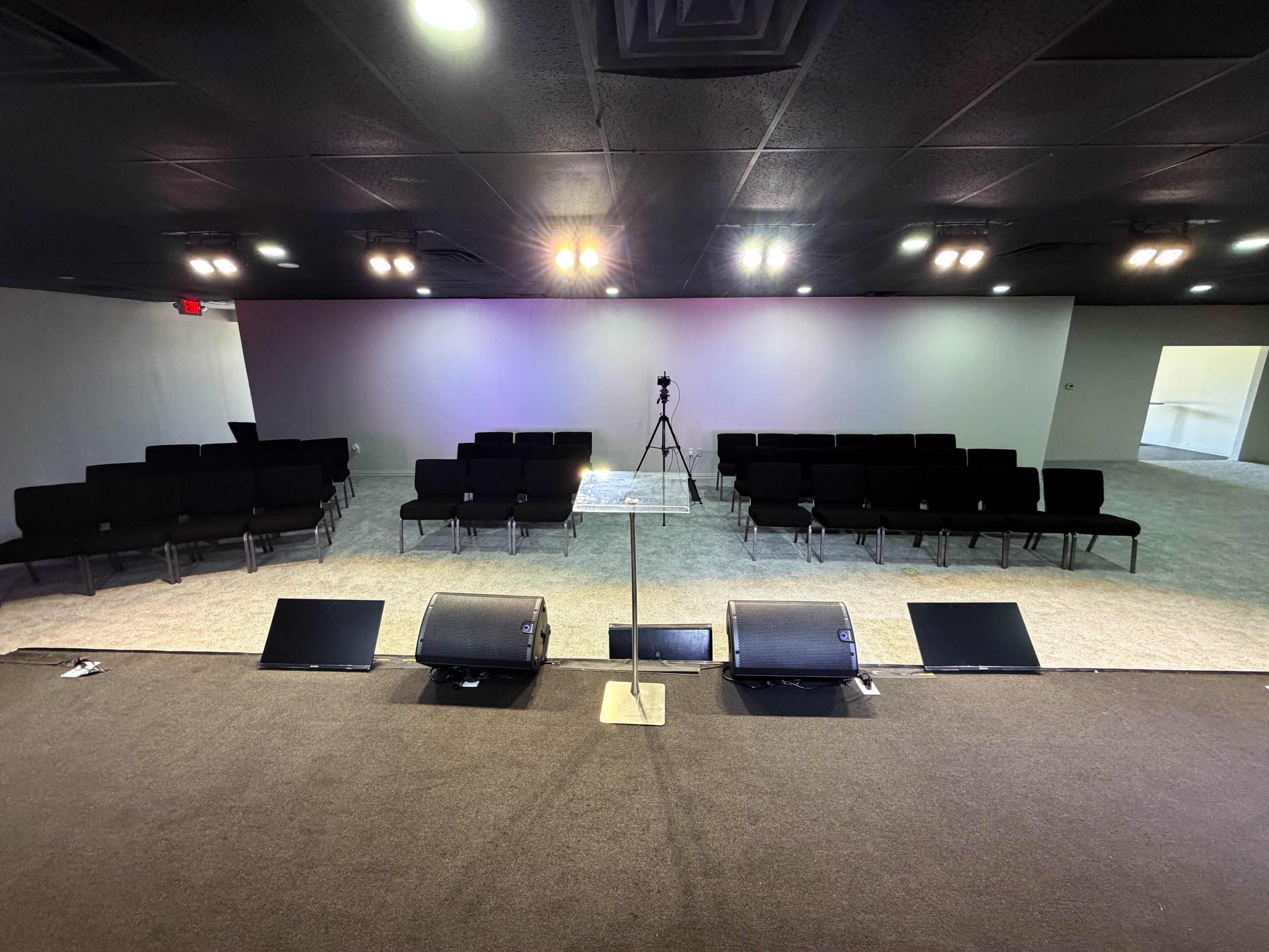 The image shows an interior space set up for a presentation or gathering, featuring rows of black chairs facing a raised platform with audio equipment and a camera positioned in the center.