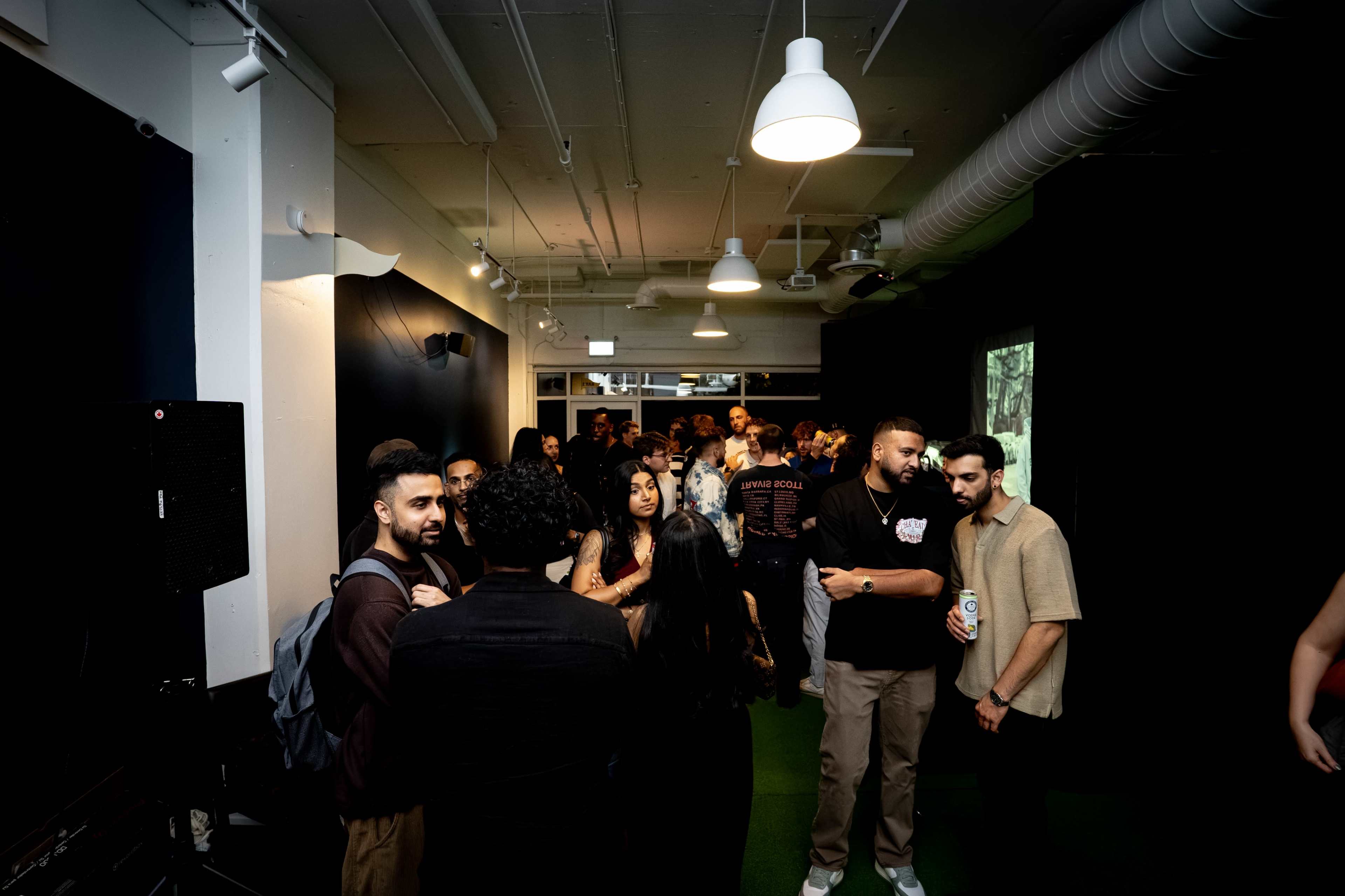 A group of people socialize in a dimly lit room with dark walls and an illuminated screen in the background.