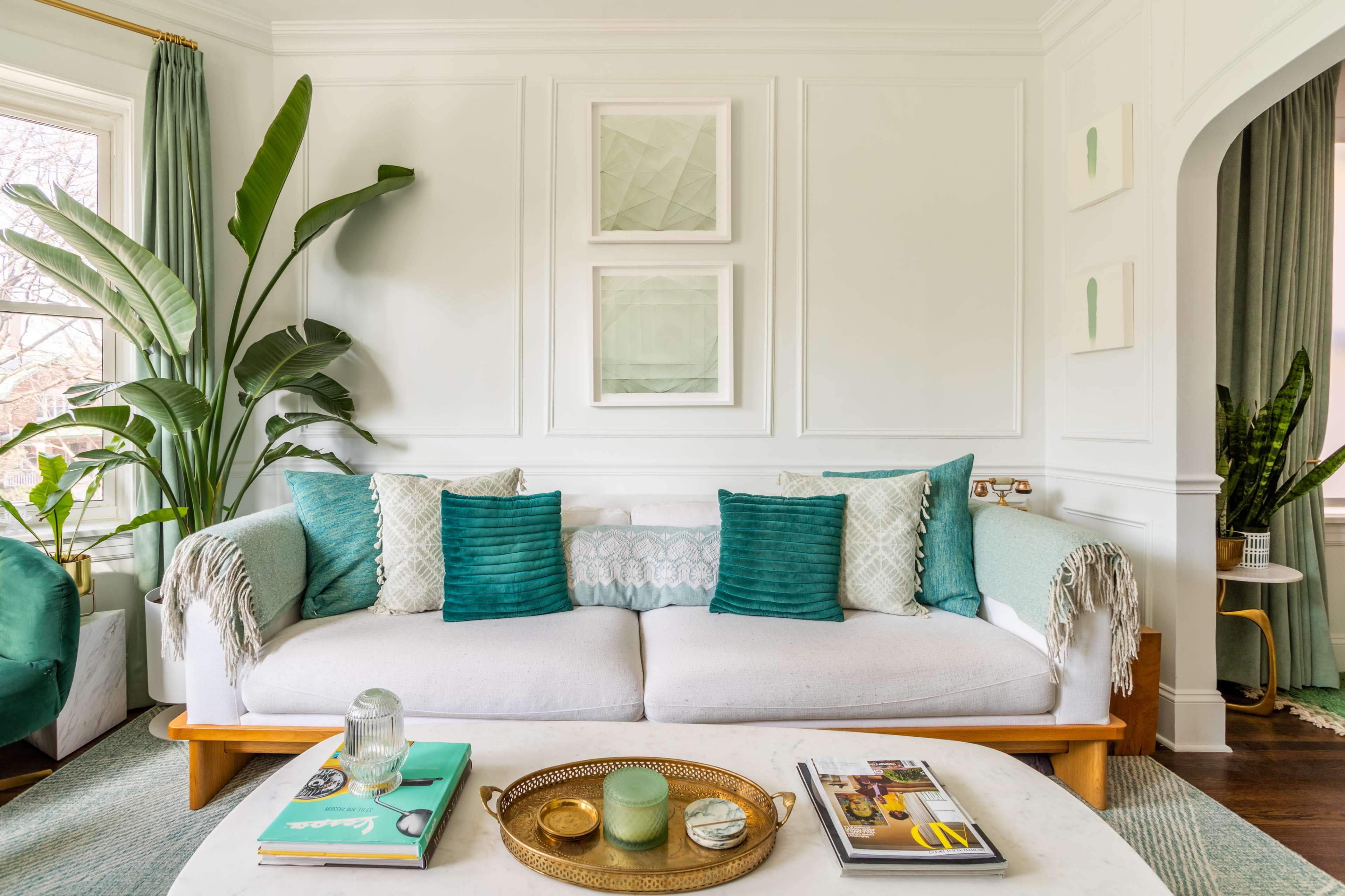 A bright living room features a white sofa adorned with teal pillows, surrounded by green plants and decorative art on the walls.