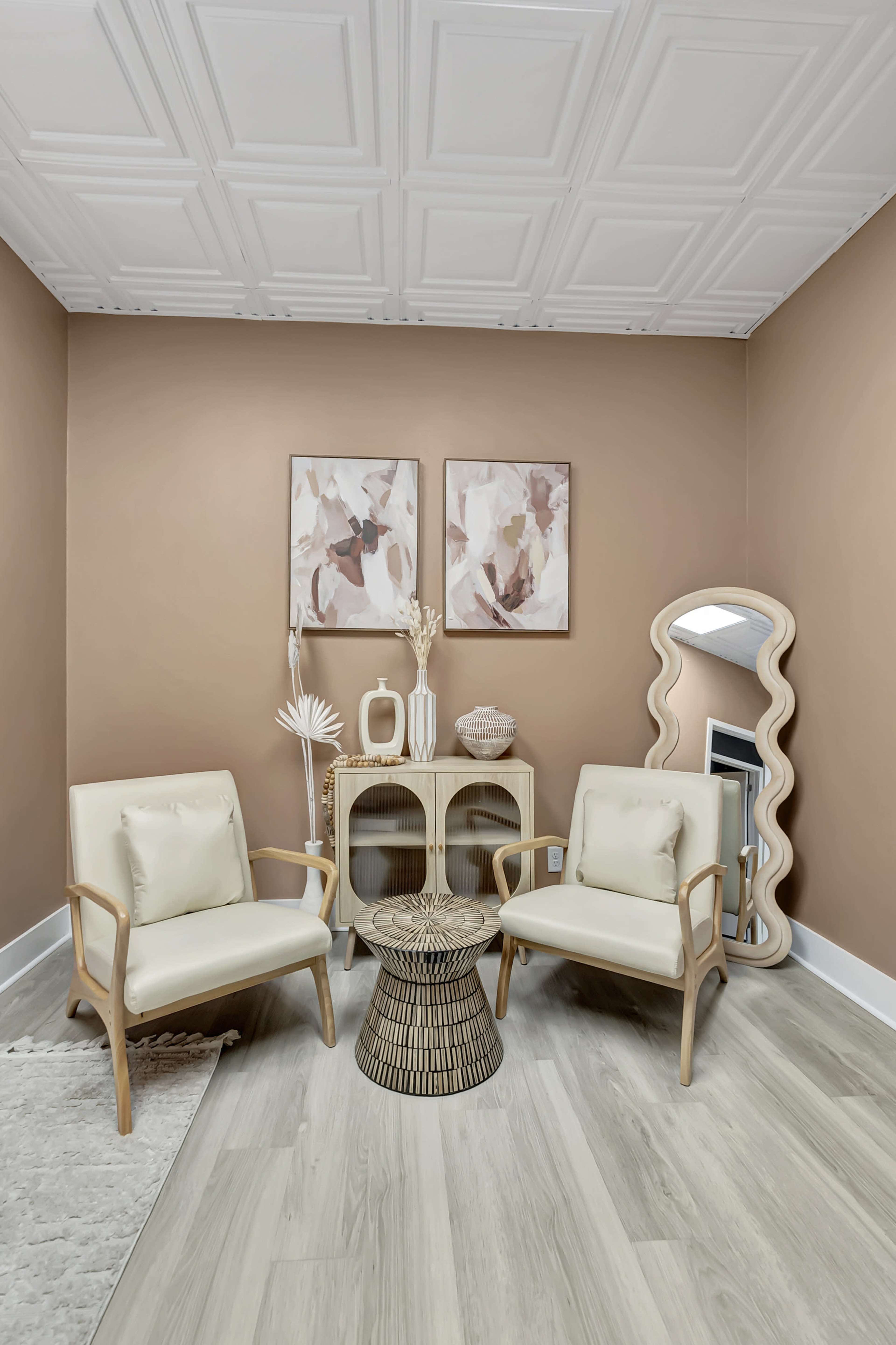 A cozy seating area features two light-colored chairs, a woven stool, and a small table, with framed artwork and a mirror against a tan wall.