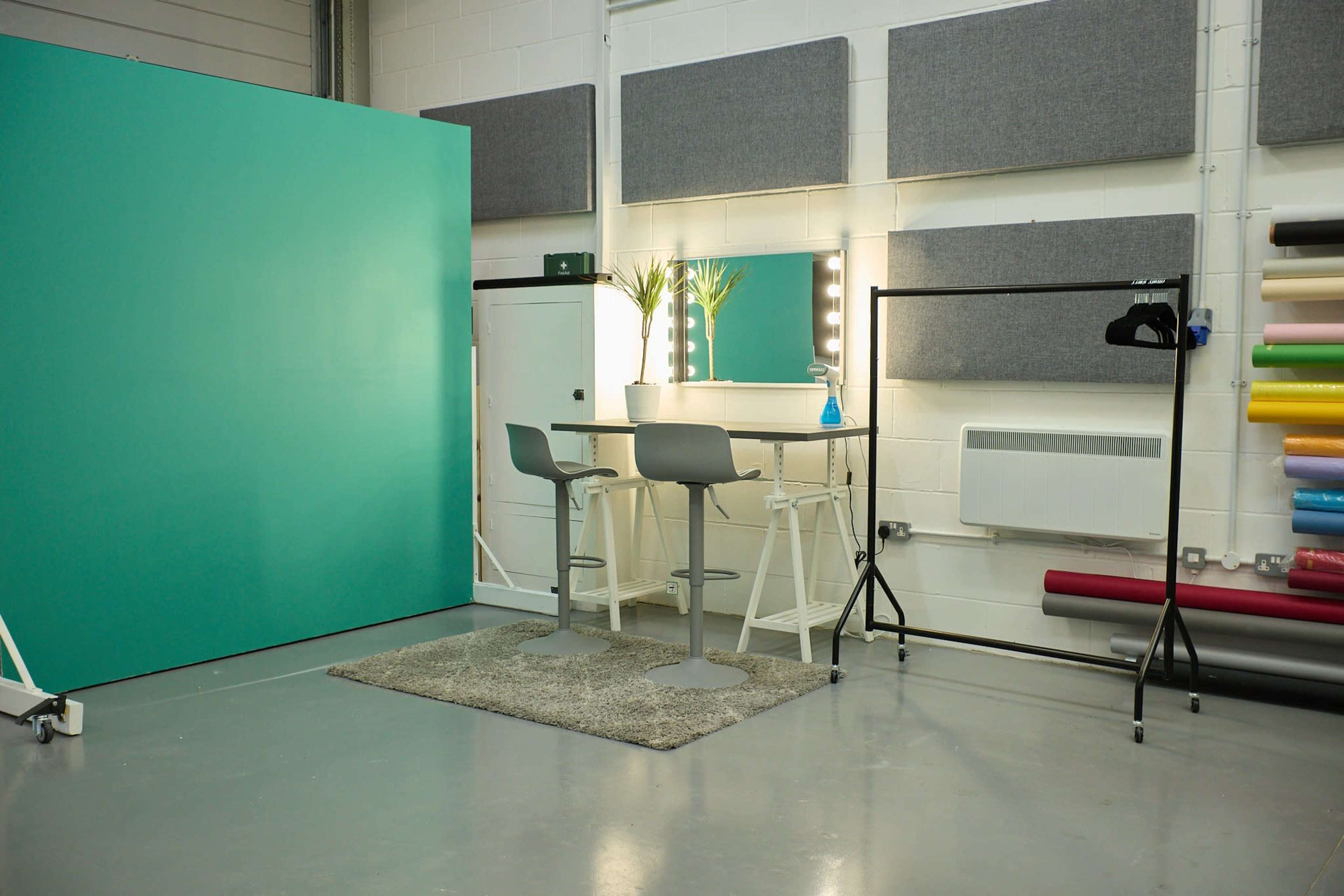 A minimalistic studio space featuring a teal backdrop, a pair of bar stools at a table with a mirror and lights, and a rolling clothing rack against a wall of acoustic panels.