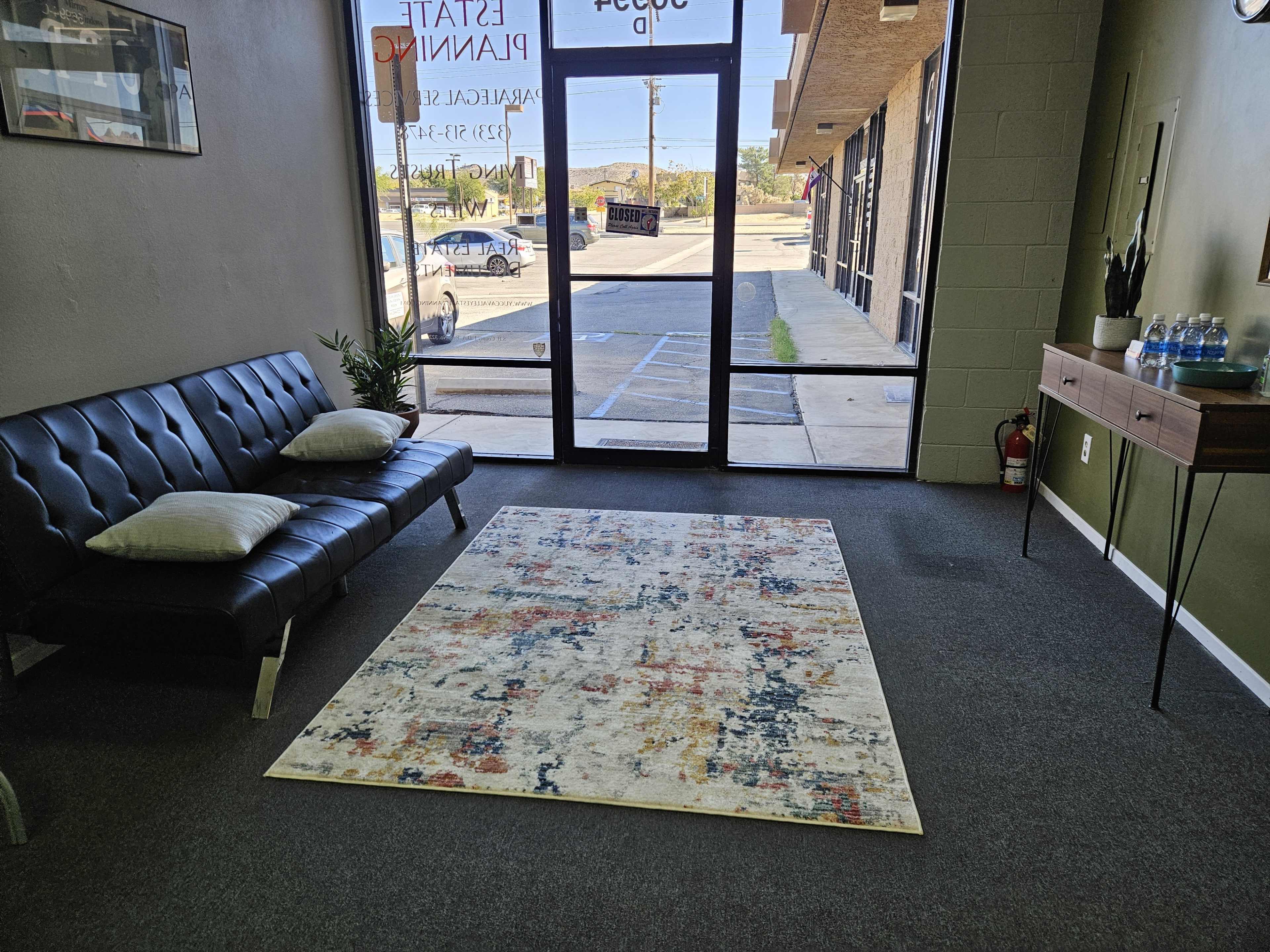 A waiting area with a black sofa, a patterned rug on the carpeted floor, and a view of a parking lot through large glass doors.