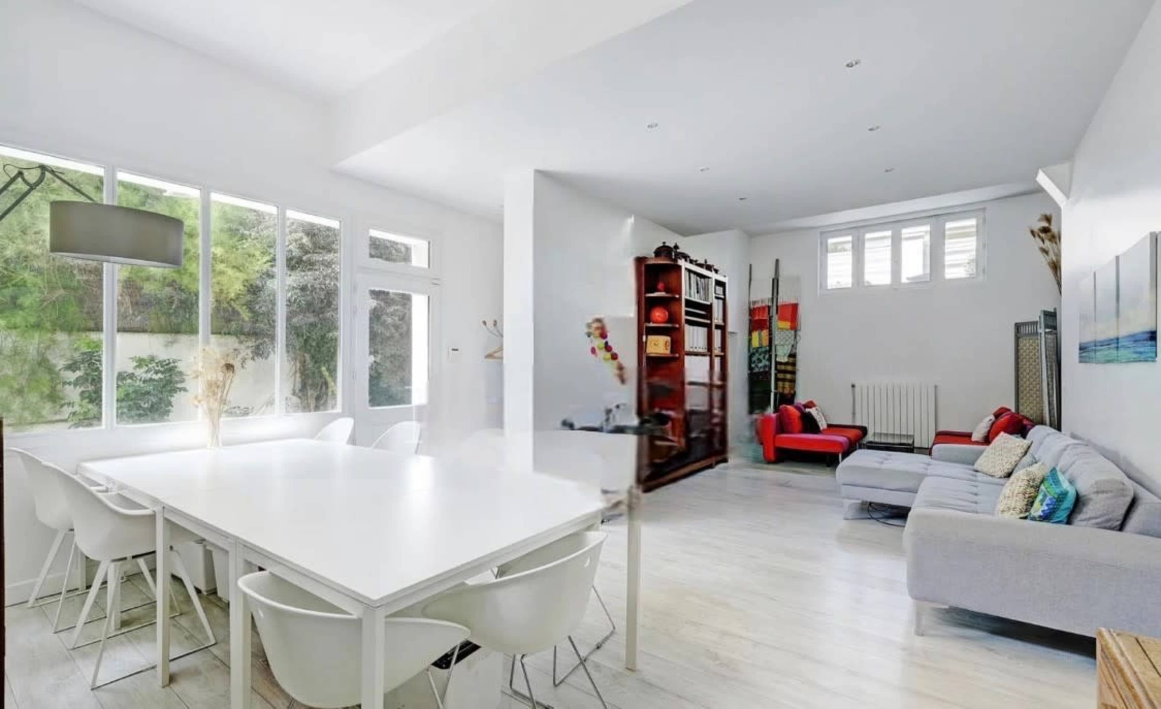 The image shows a bright, modern living space with a large dining table, white chairs, and a cozy seating area featuring a gray sofa and a red armchair.