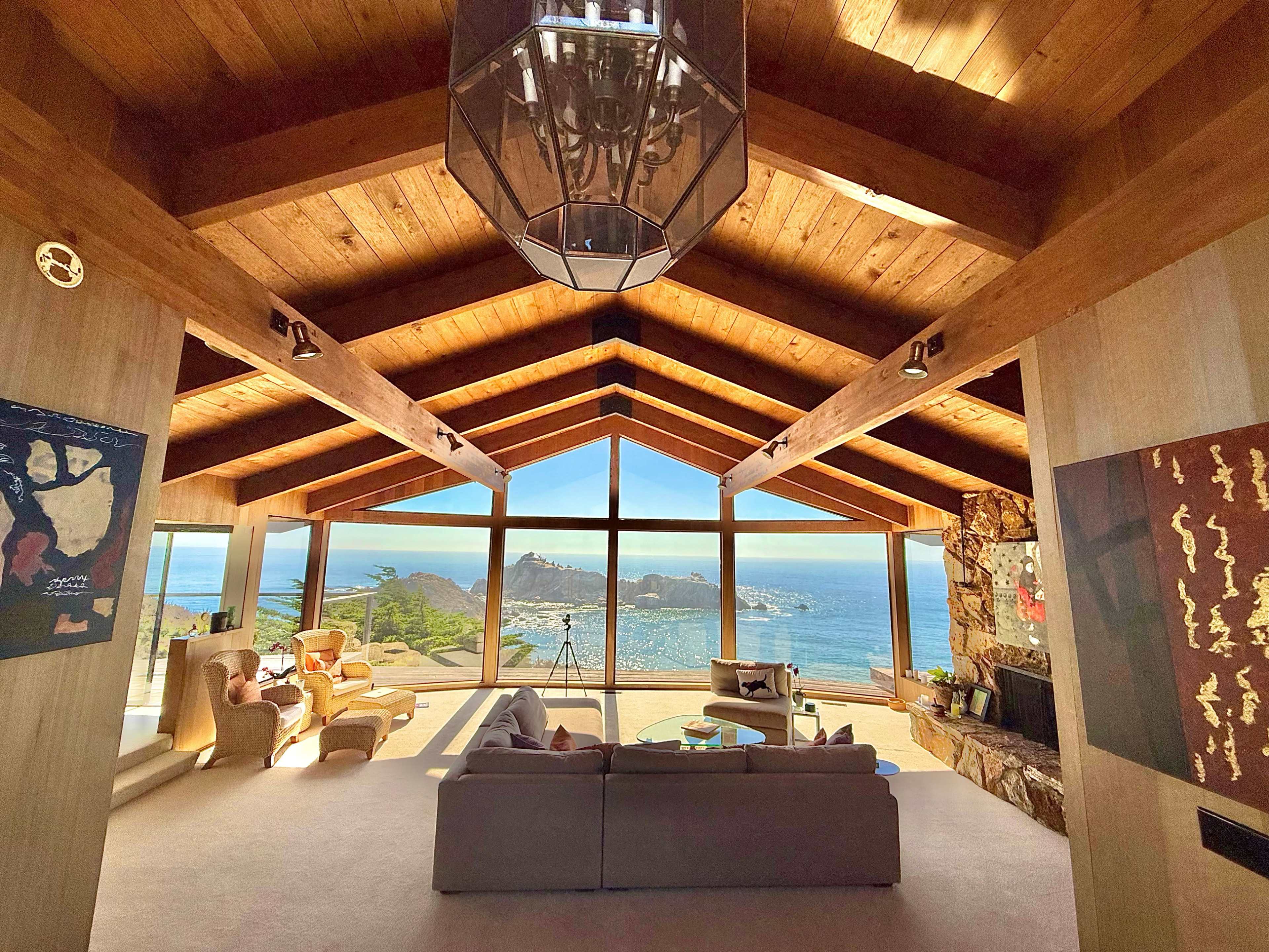 The image shows a spacious living room with a high wooden ceiling, large windows overlooking a coastal view, and modern furniture arranged invitingly.