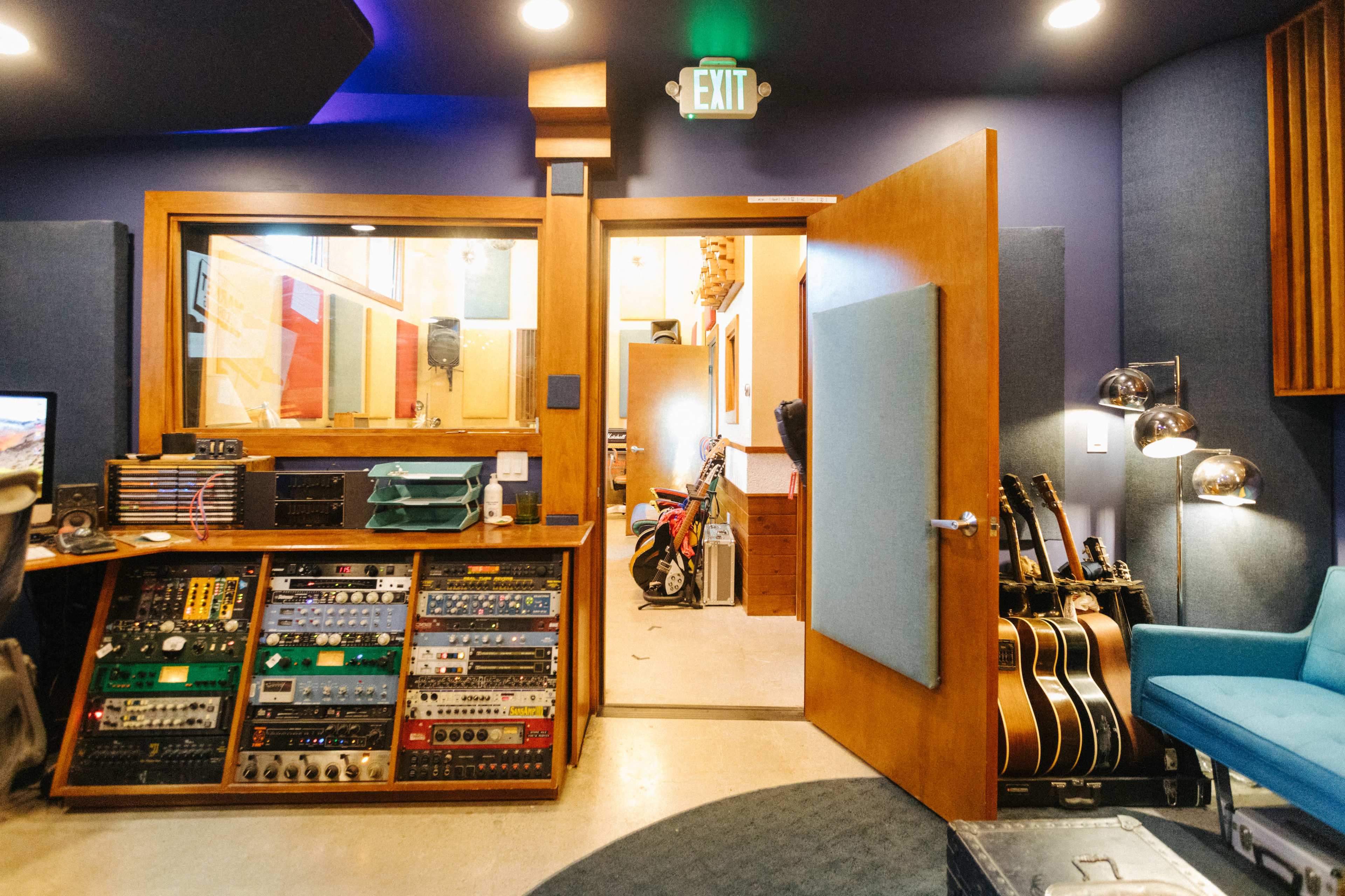 A music studio with audio equipment on a console, an open door leading to another room, and guitars leaning against the wall.