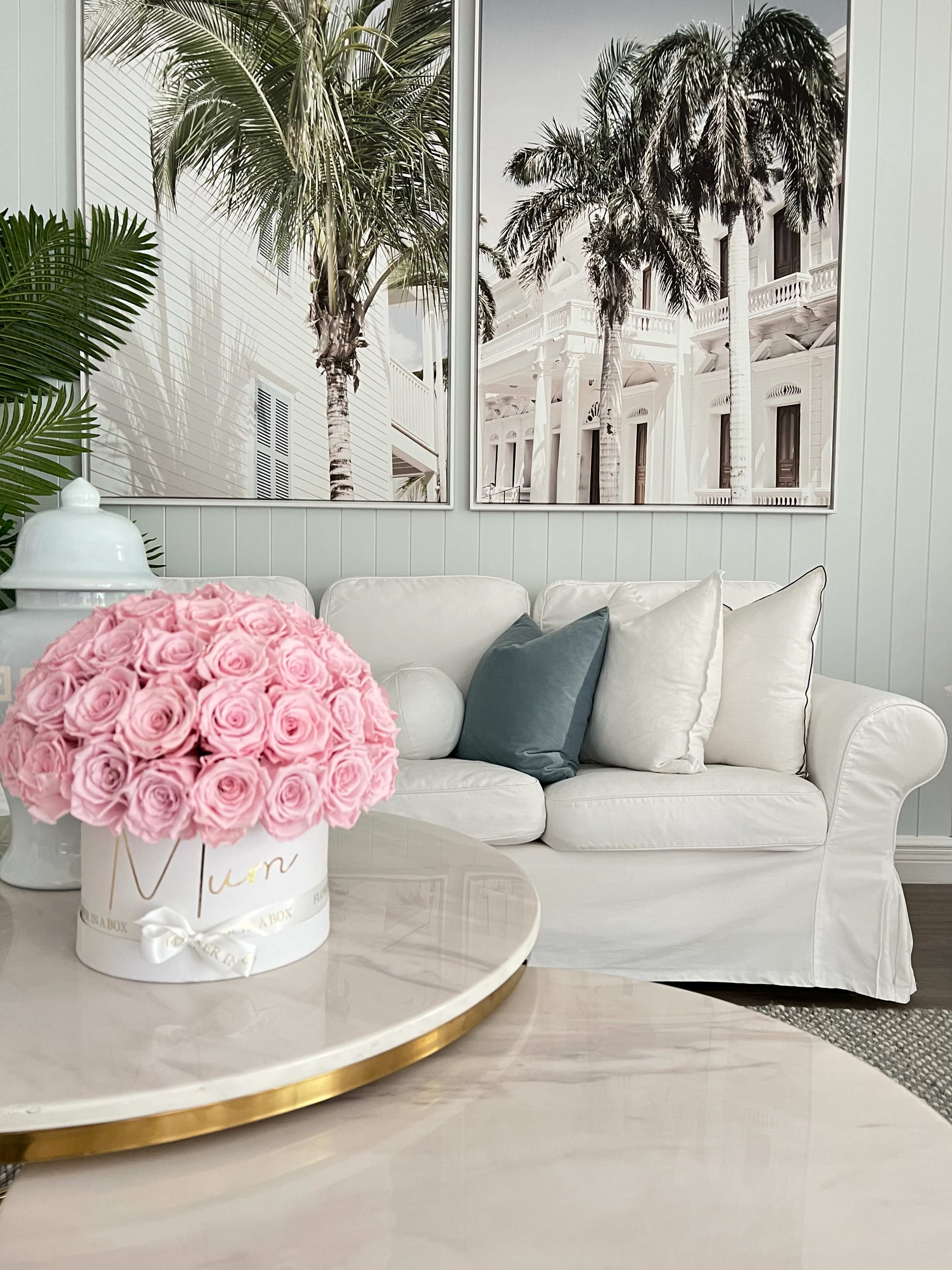 A living room features a white couch adorned with pillows, a marble coffee table, and a round box of pink roses set beside framed palm tree photographs.