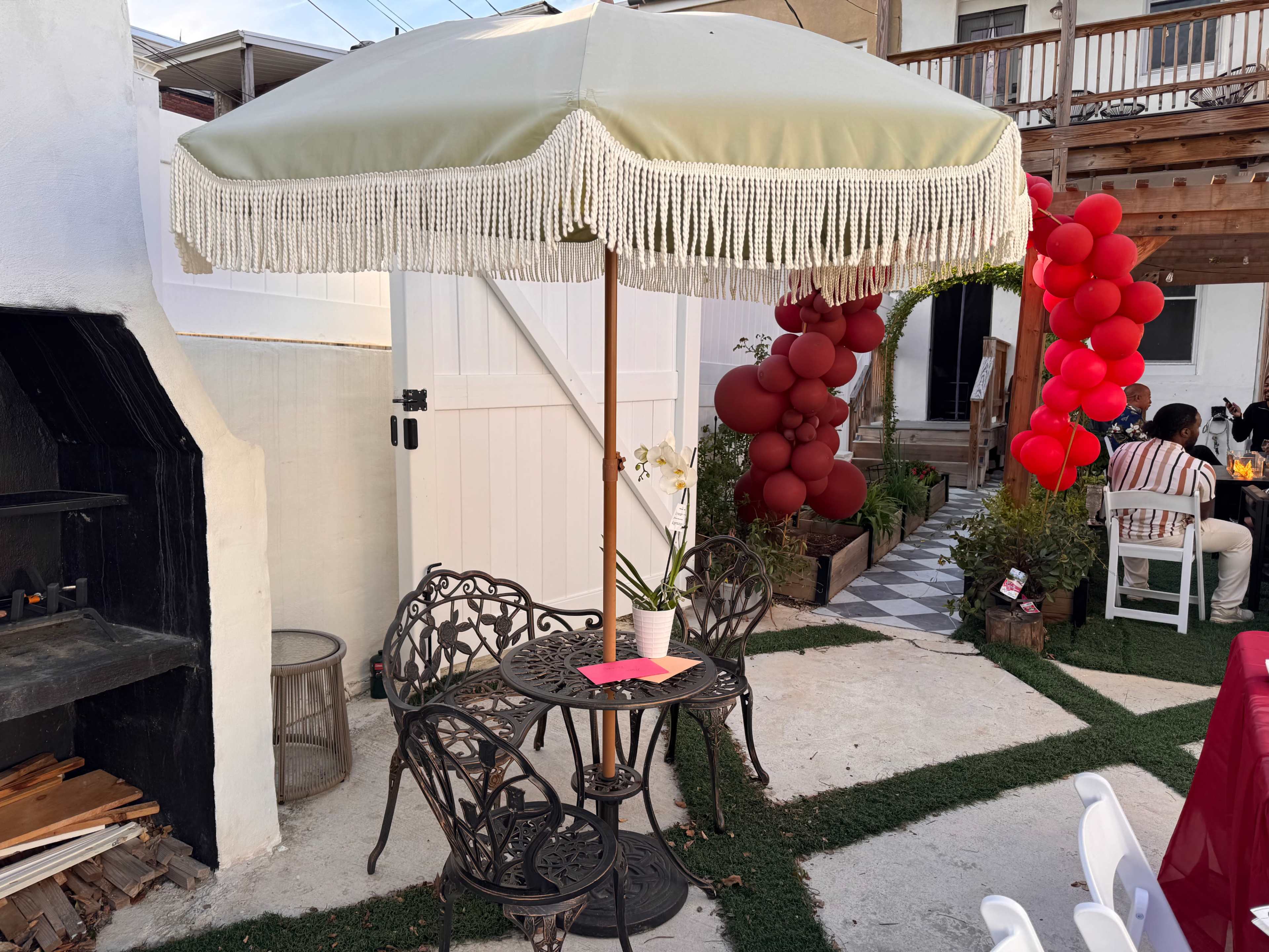 A small outdoor seating area features a round table and two chairs under a fringed umbrella, with red balloons and party decorations nearby.