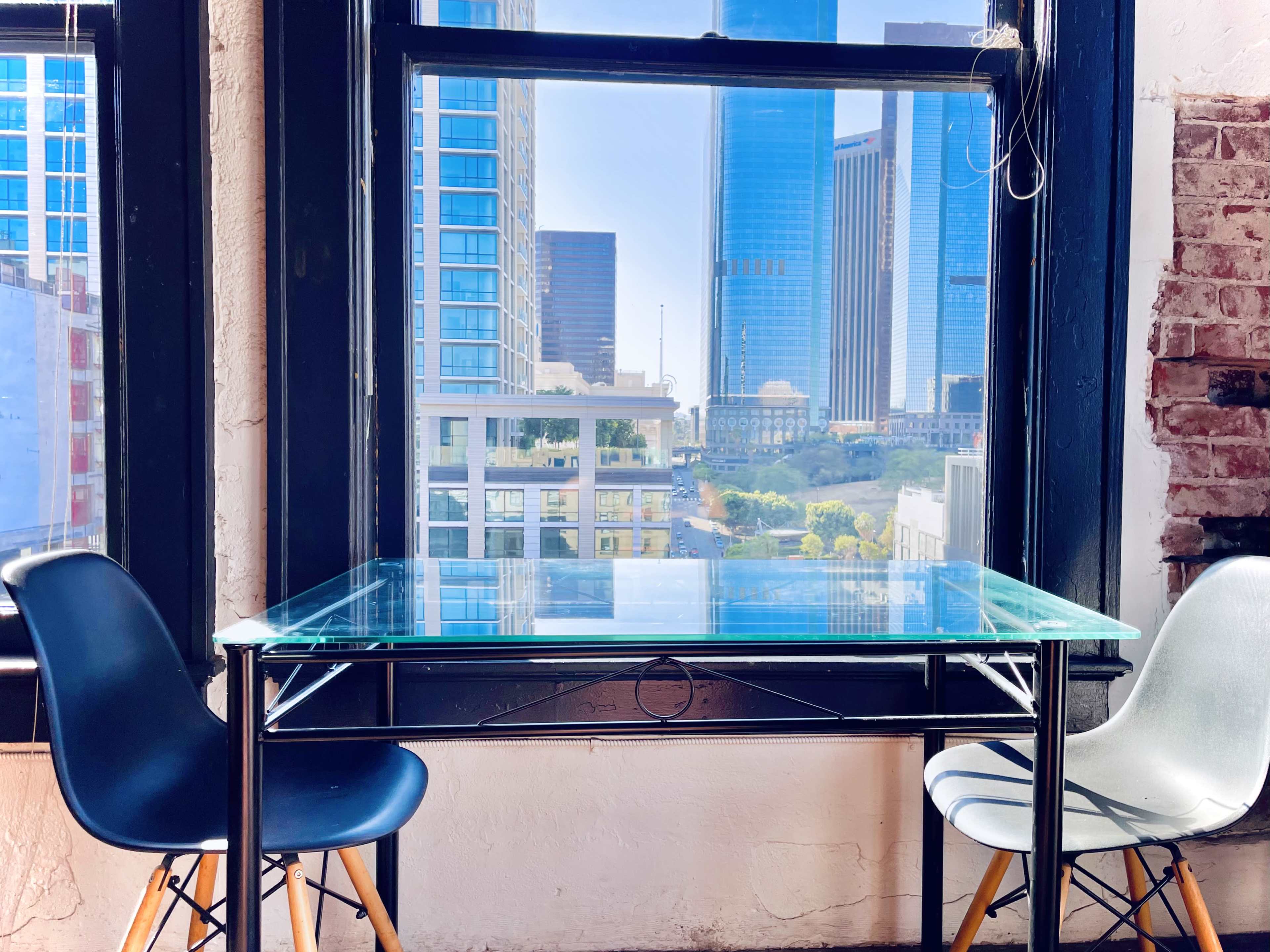 A glass table with black and white chairs sits in front of a large window overlooking a city skyline with tall buildings.