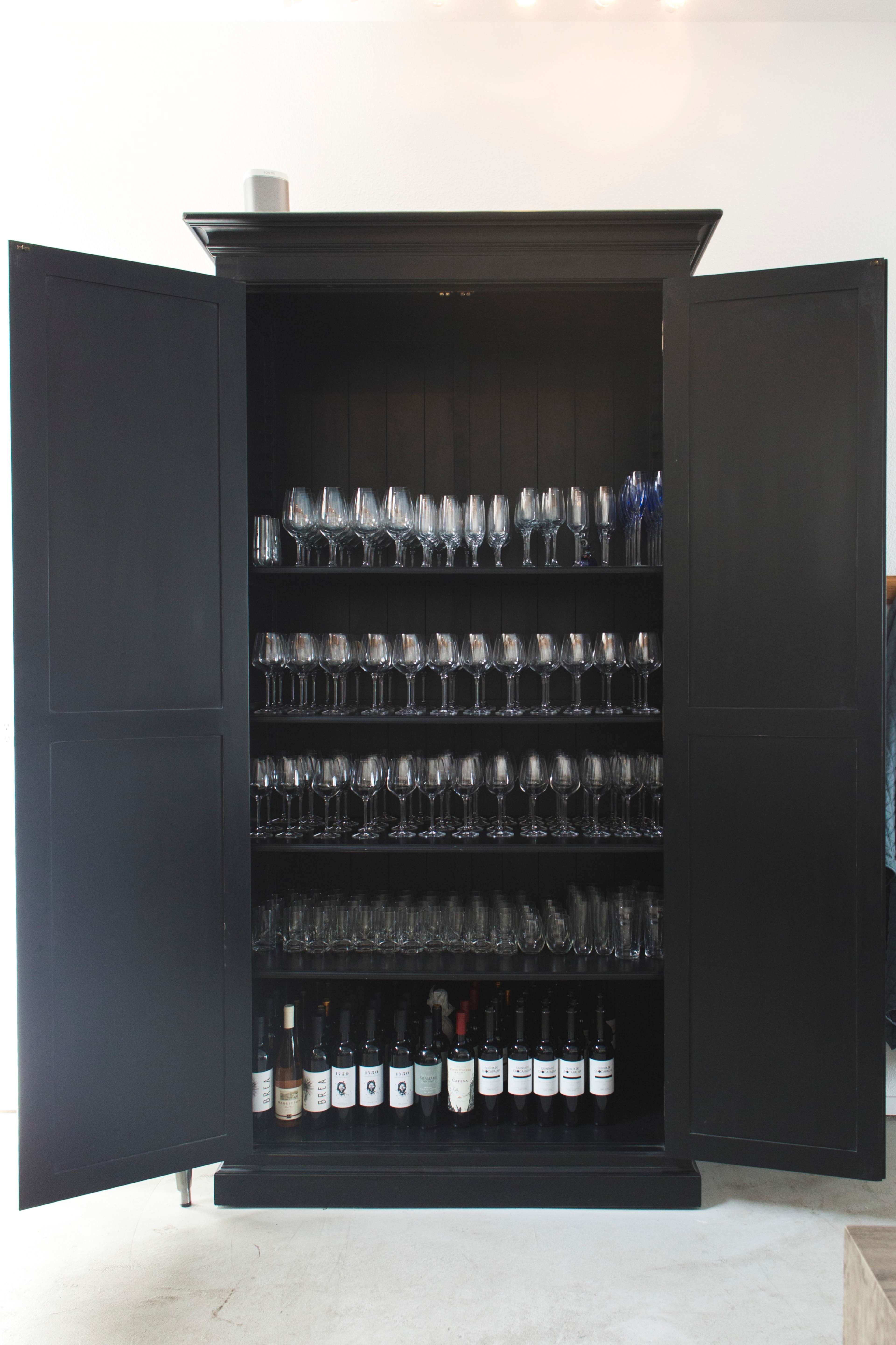 A black cabinet with open double doors displays several rows of glassware and bottles of wine.