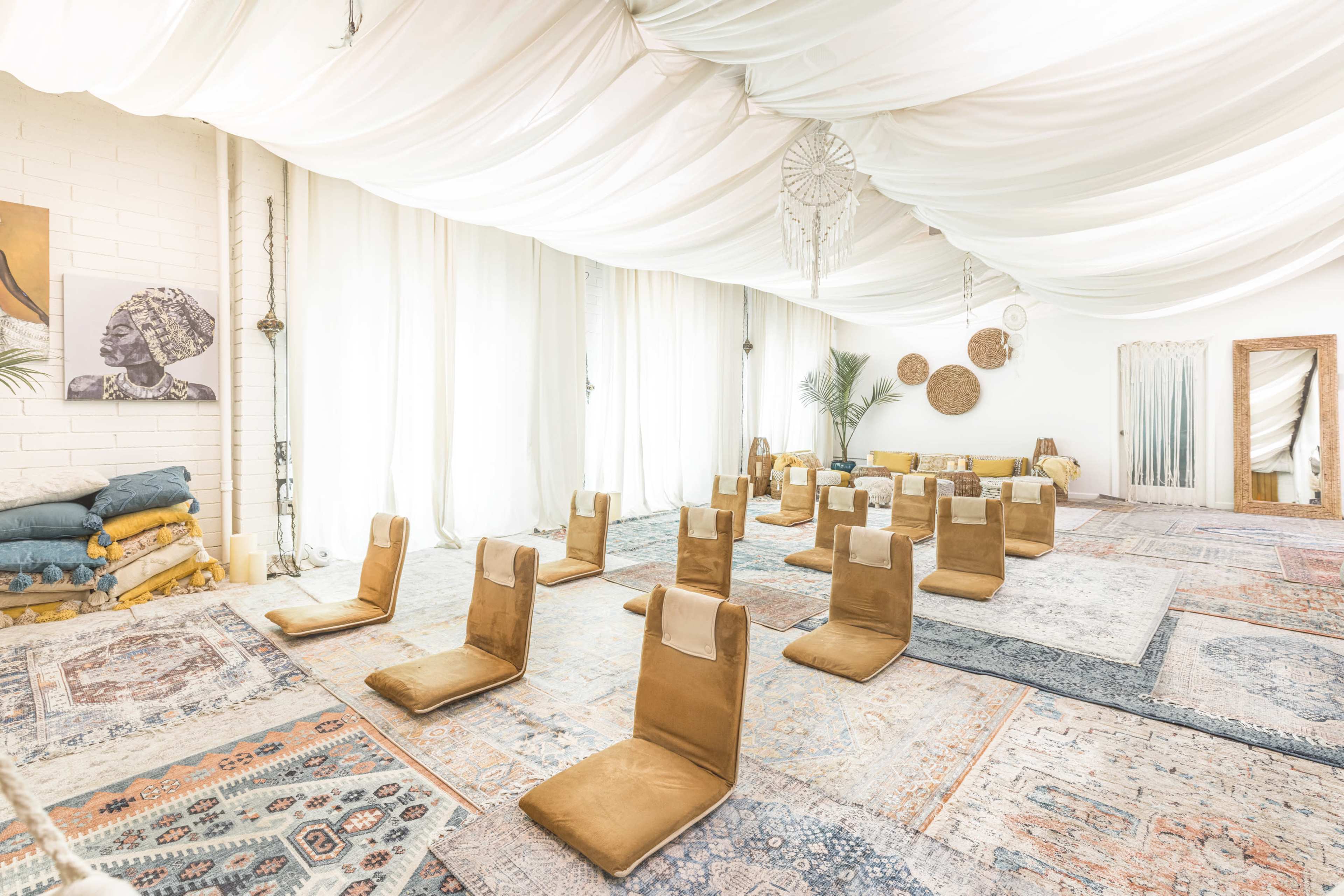 A spacious, well-lit room arranged with several floor seating pads on intricately patterned rugs, surrounded by light-colored drapes and decorative wall hangings.
