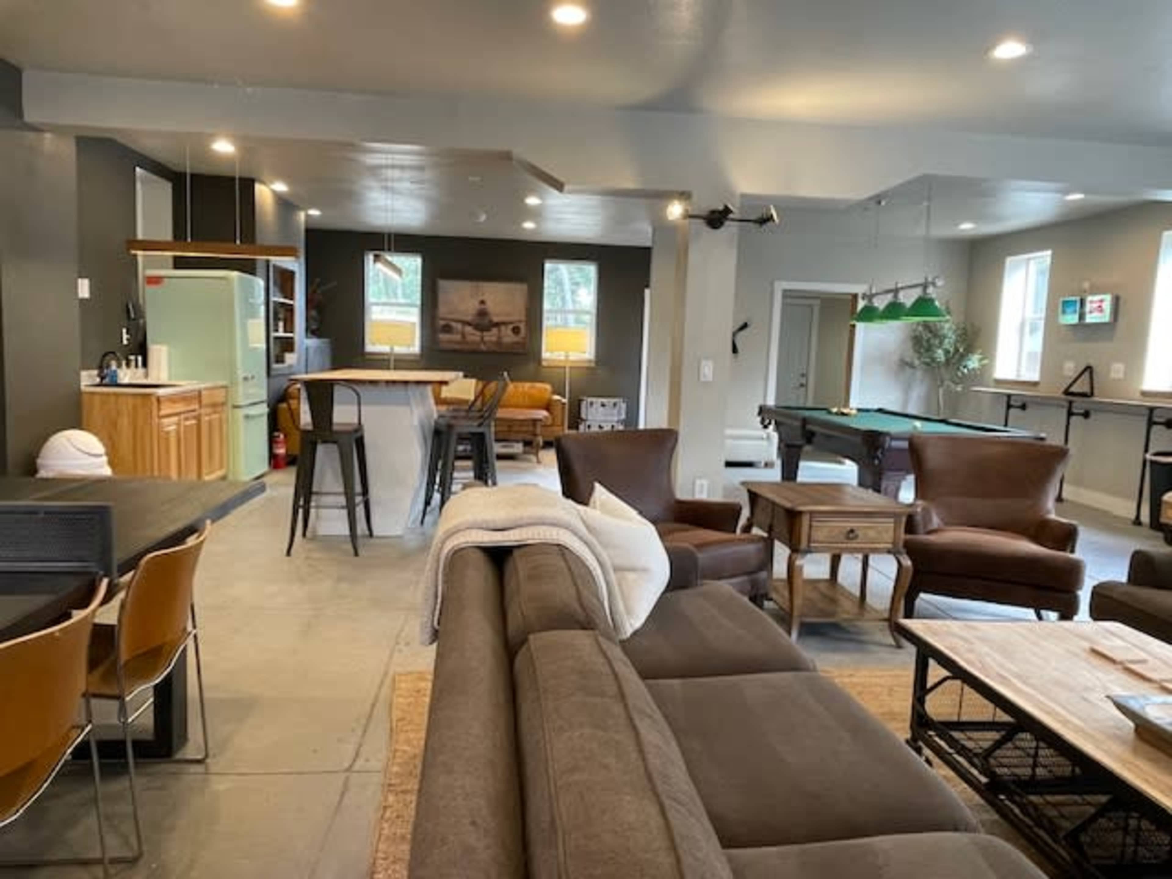 A spacious, modern living area featuring a mix of seating options, a kitchen area, a billiard table, and large windows allowing natural light.