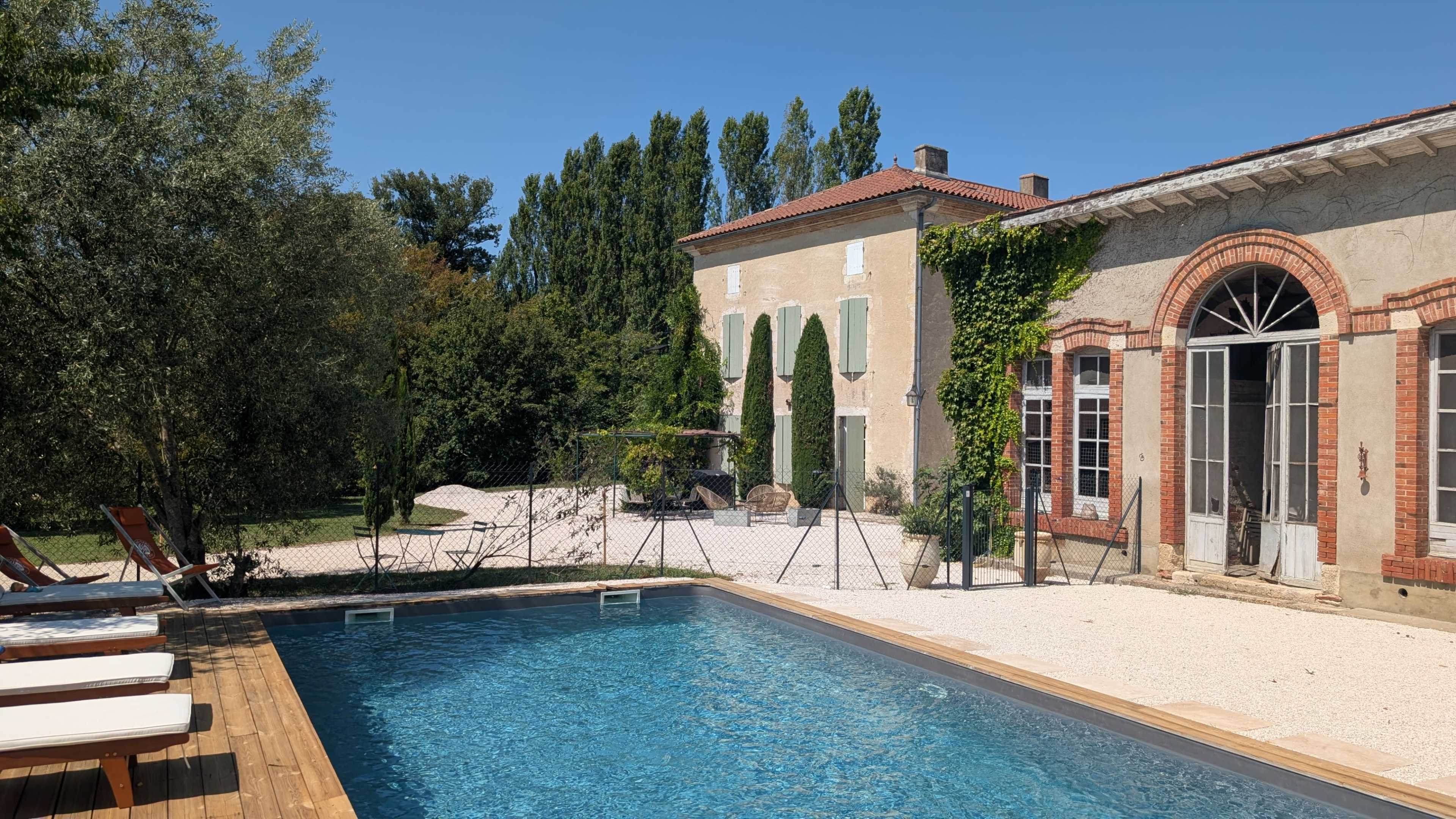 The image shows a swimming pool with wooden decking in the foreground, surrounded by a landscaped garden and a large rustic building in the background.