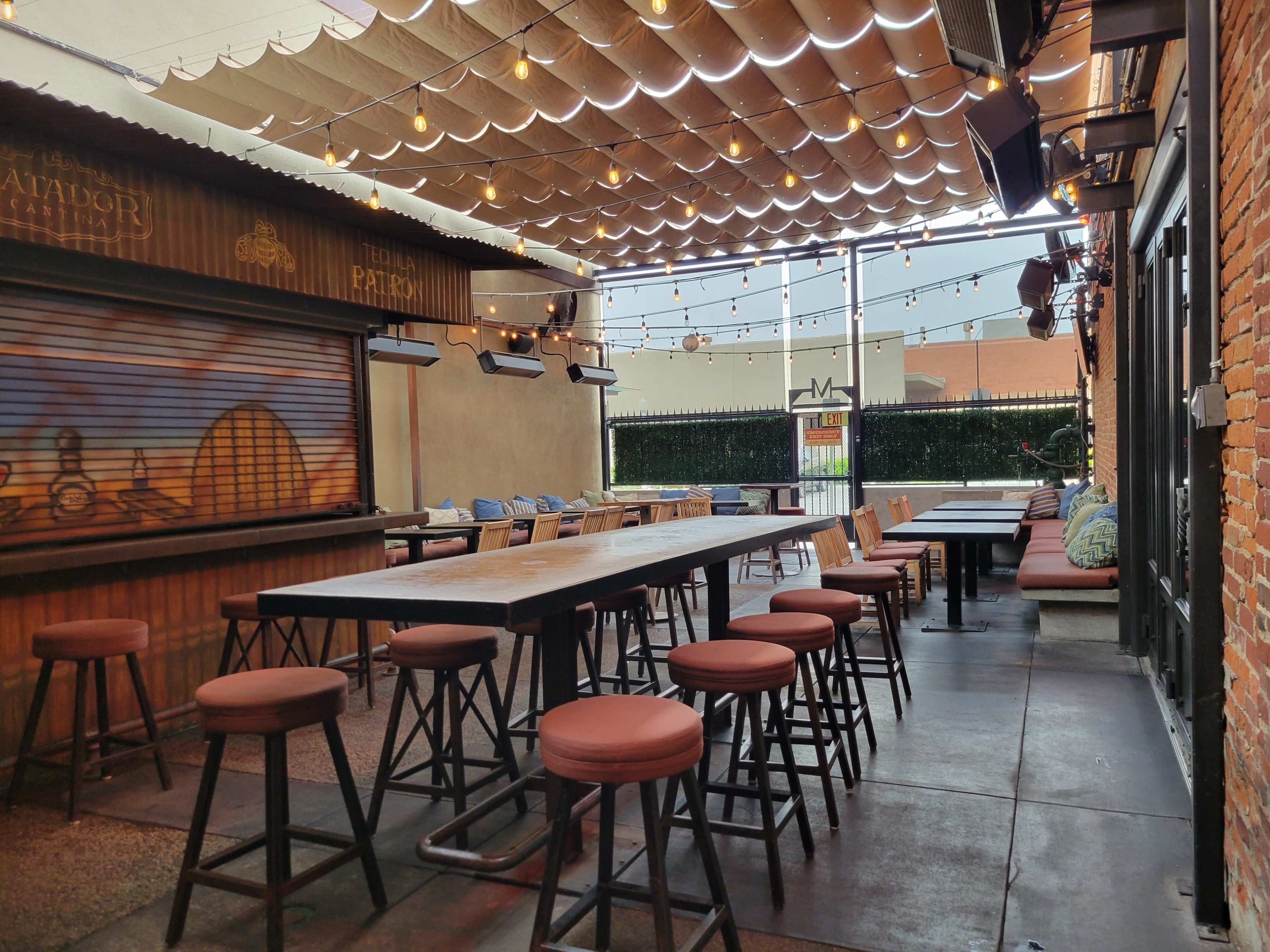 An outdoor restaurant patio featuring wooden tables, high stools, and string lights overhead.