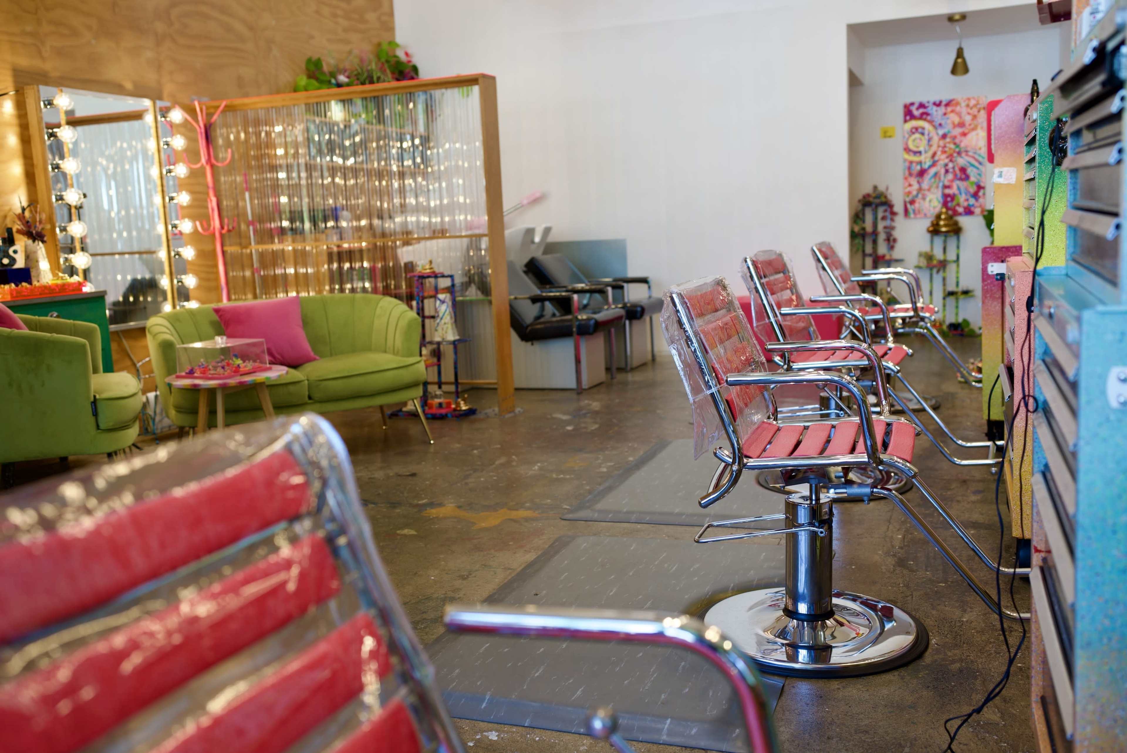 The image shows a modern hair salon featuring chrome salon chairs with red cushions, a green couch, and decorative elements in a spacious, well-lit interior.