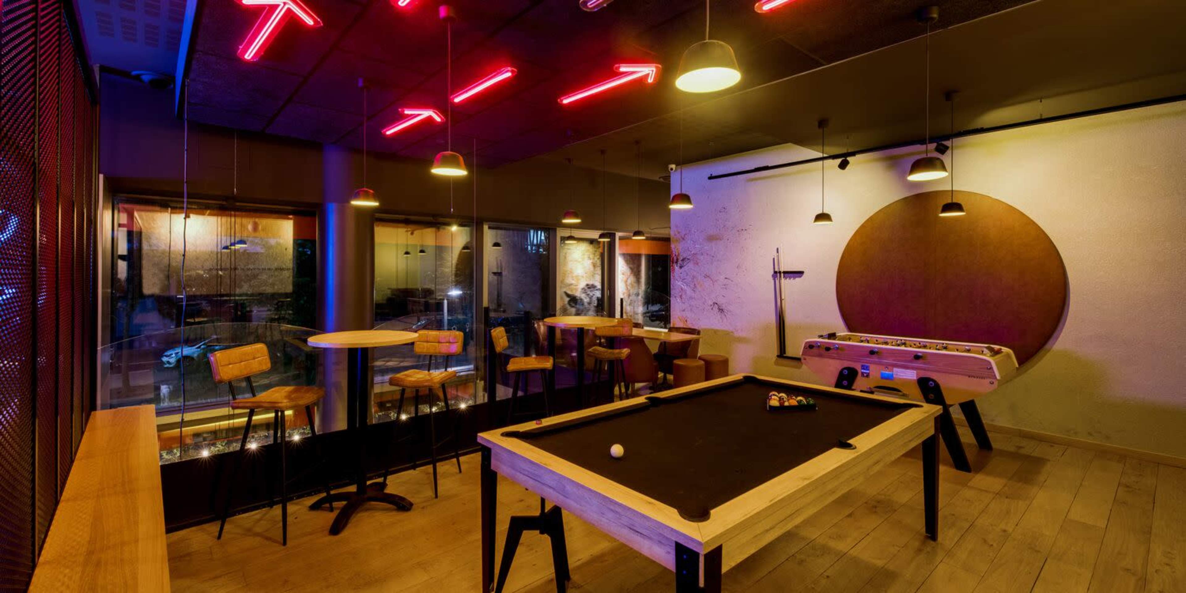 A game room with a pool table, foosball table, and neon lighting, featuring wooden stools and tables against a backdrop of large windows.