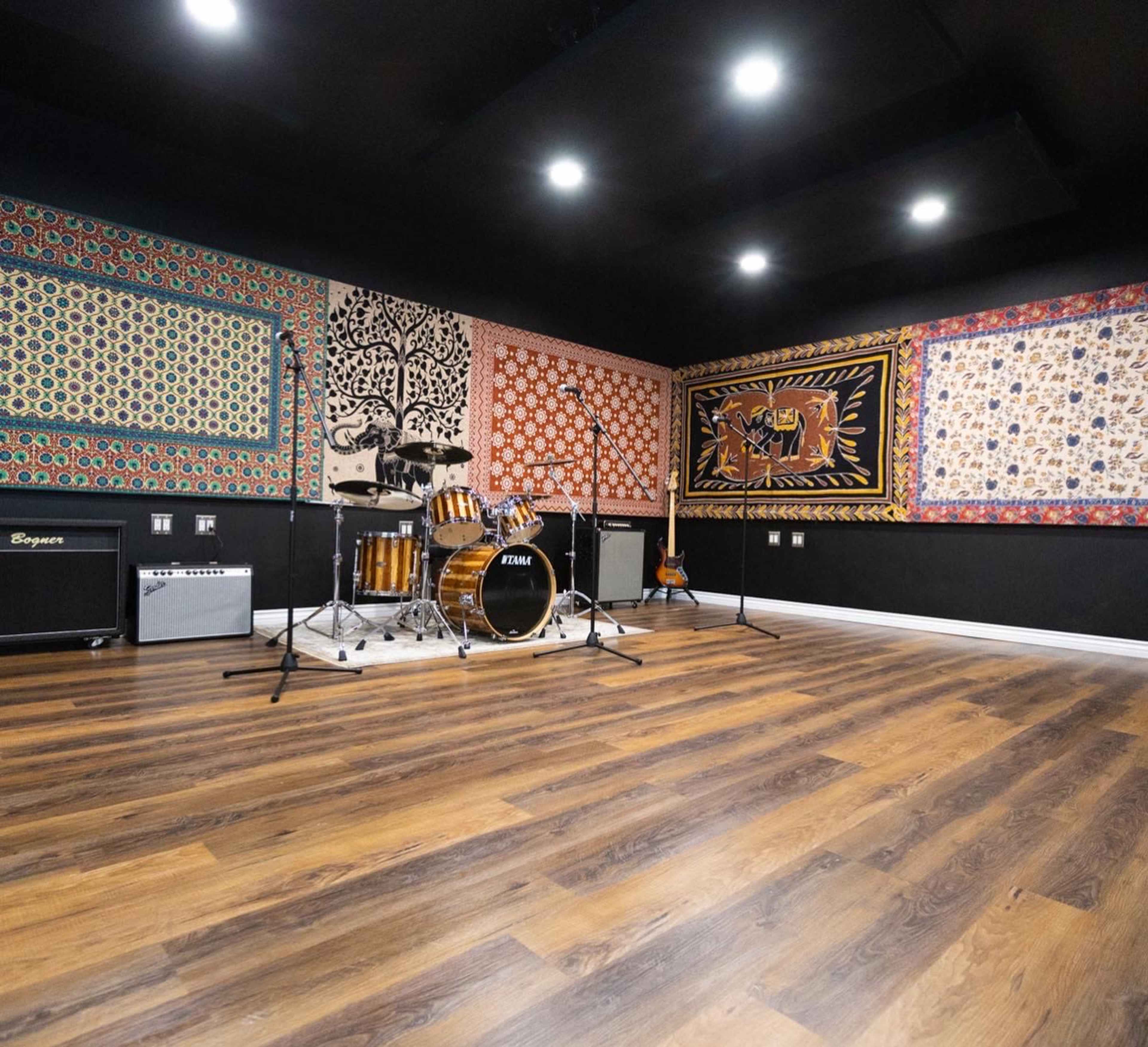 The image shows a music studio with a drum set, electric guitar, and amplifiers arranged against walls adorned with colorful patterned tapestries.