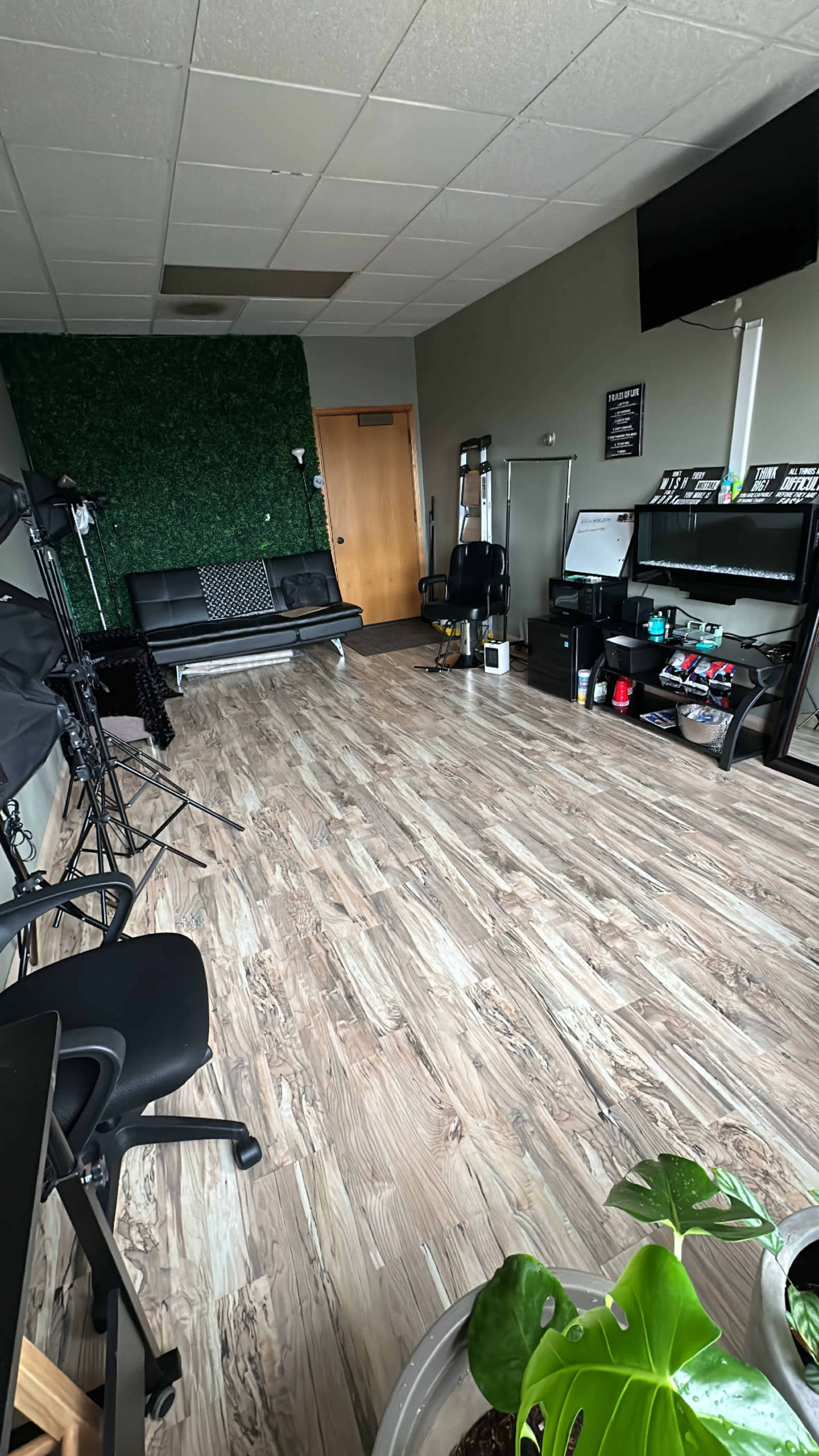 The image shows a spacious room with wooden flooring, featuring a black sofa, a desk with various items, and decorative green wall plants.