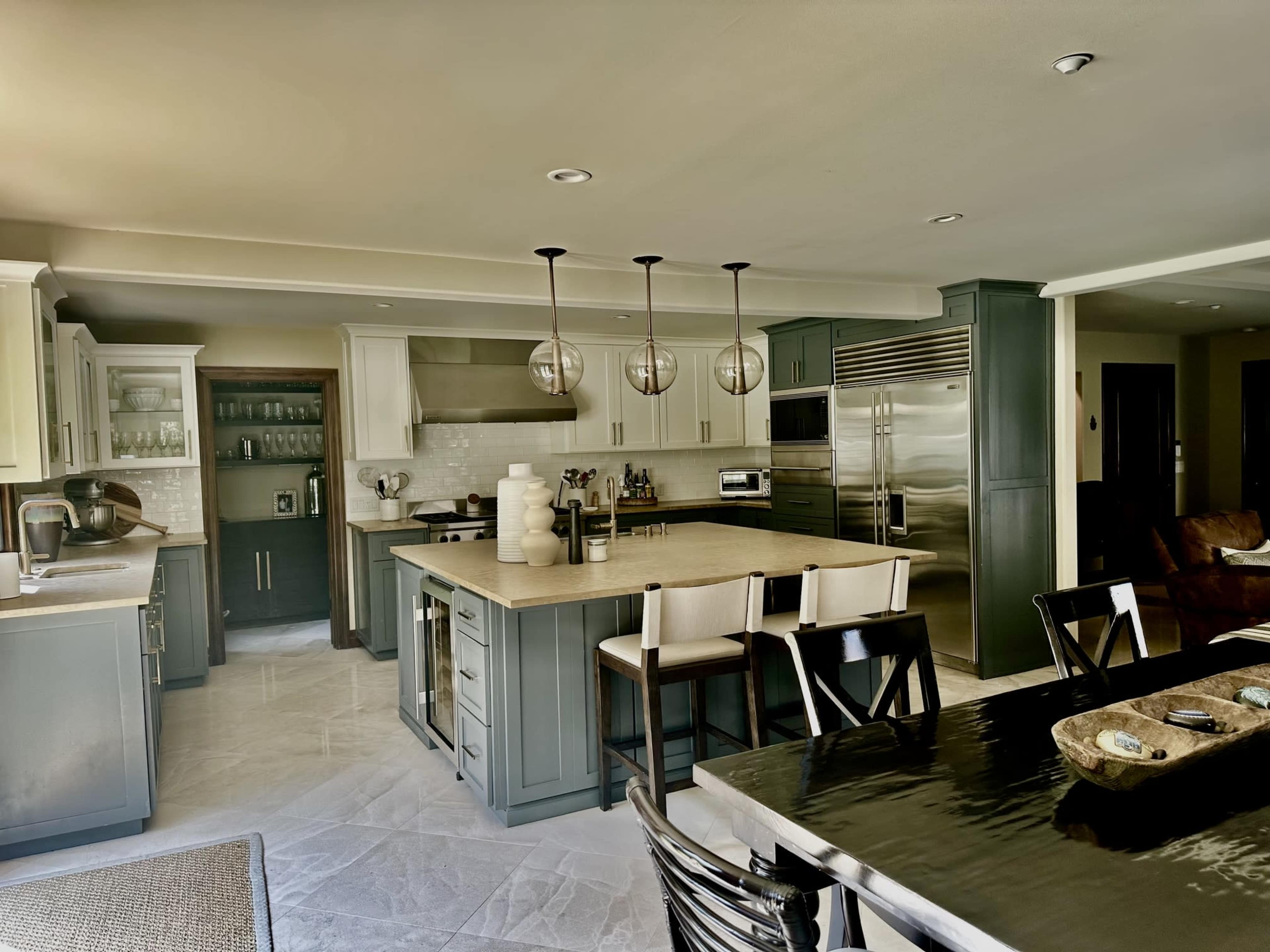 The image shows a modern kitchen featuring gray cabinetry, stainless steel appliances, a large island with bar seating, and a dining area with a dark table and chairs.