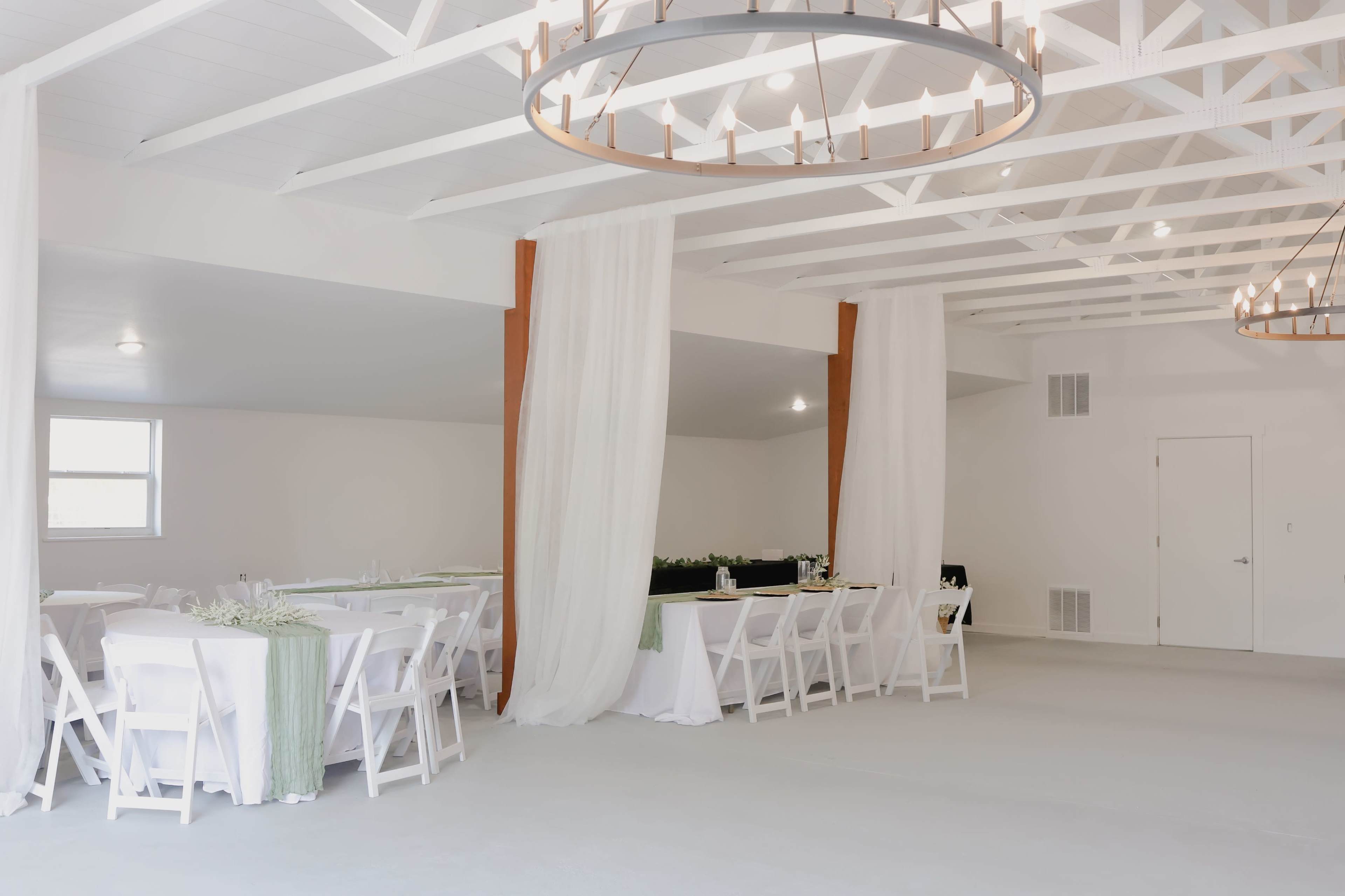 The image shows an elegantly arranged indoor event space with white tables and chairs, draped fabric, and overhead light fixtures.