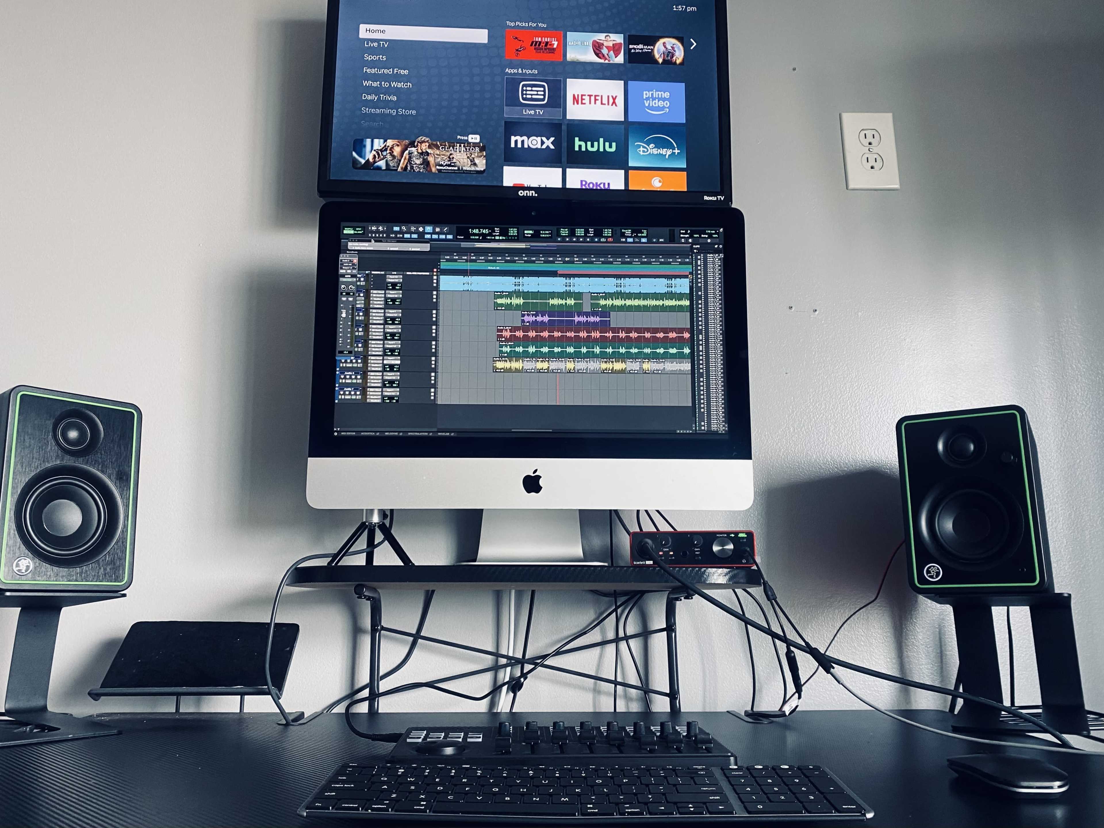 A computer workstation featuring an iMac with audio editing software displayed, flanked by studio speakers and a television mounted above showing streaming service options.