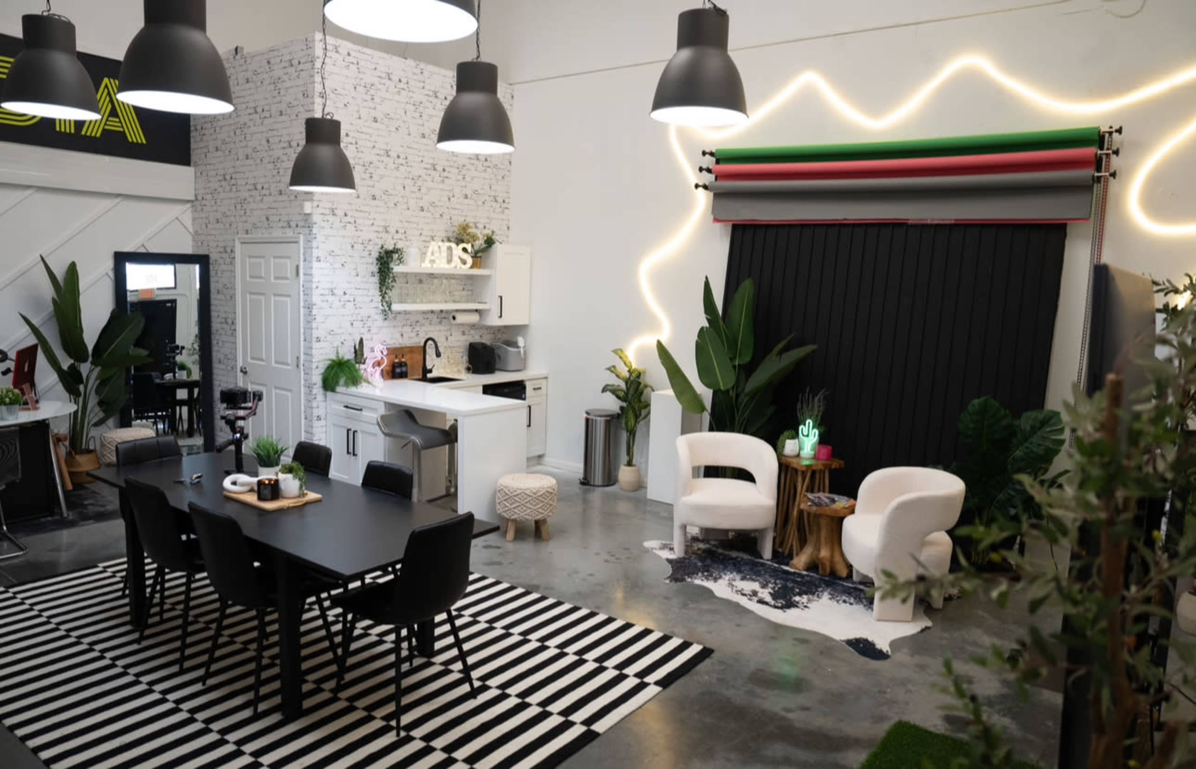 The image shows a modern studio space featuring a kitchen area, a dining table with chairs, and a seating area with decorative plants and a backdrop.